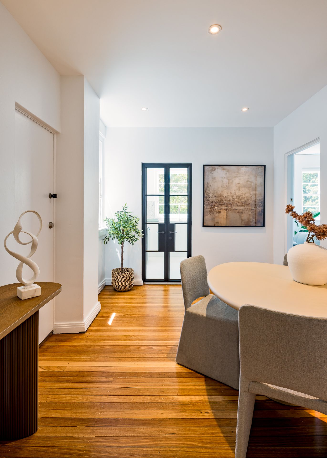Sunny dining area with round table, grey chairs, black steel French doors, potted plant and polished timber floors.