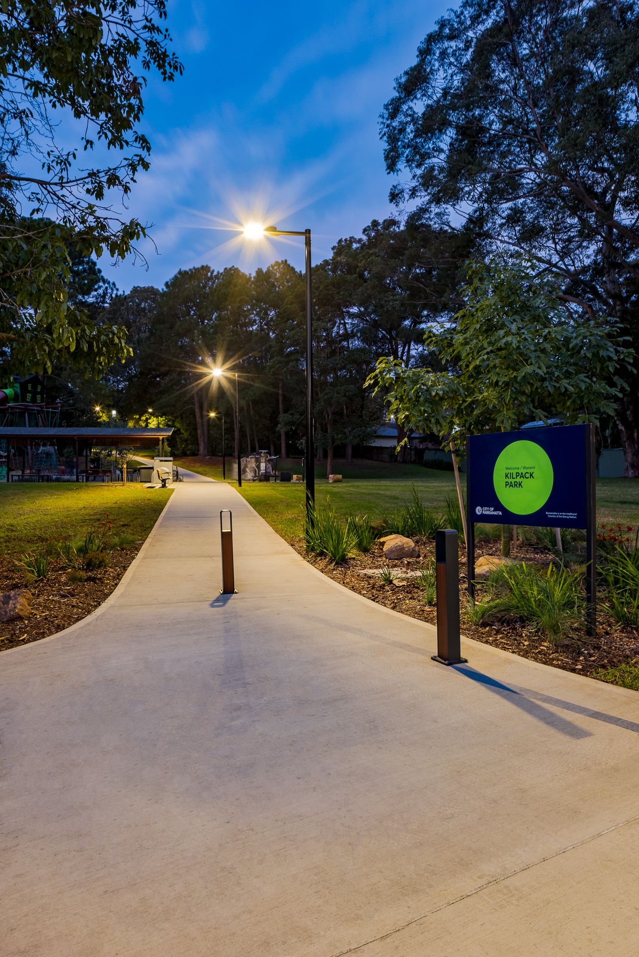 Curved concrete footpath through a landscaped park at dusk, with glowing street lamps, bollards and a green park sign among trees.