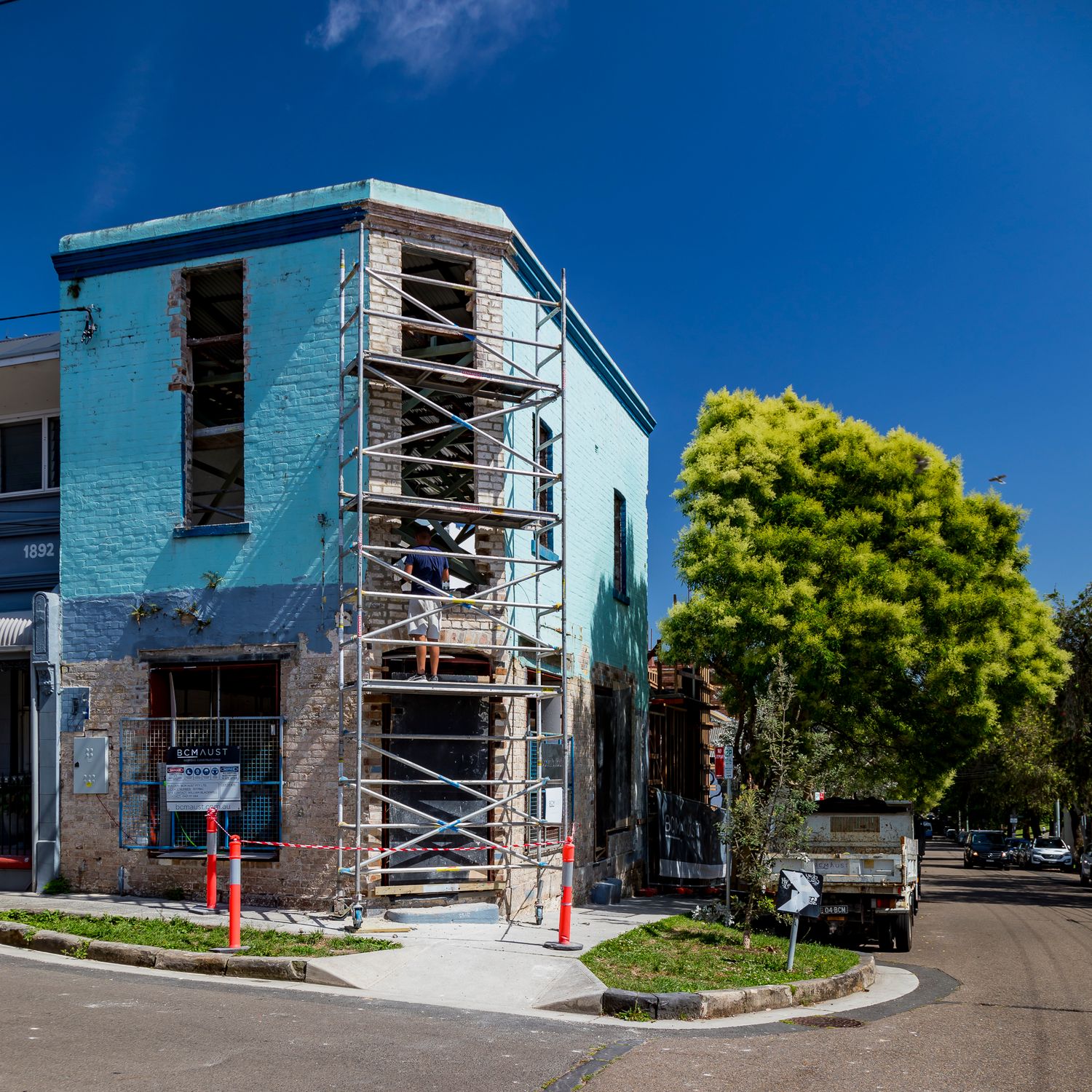 Corner brick building with blue painted facade and scaffolding during renovation, with a leafy tree and a truck on the street.