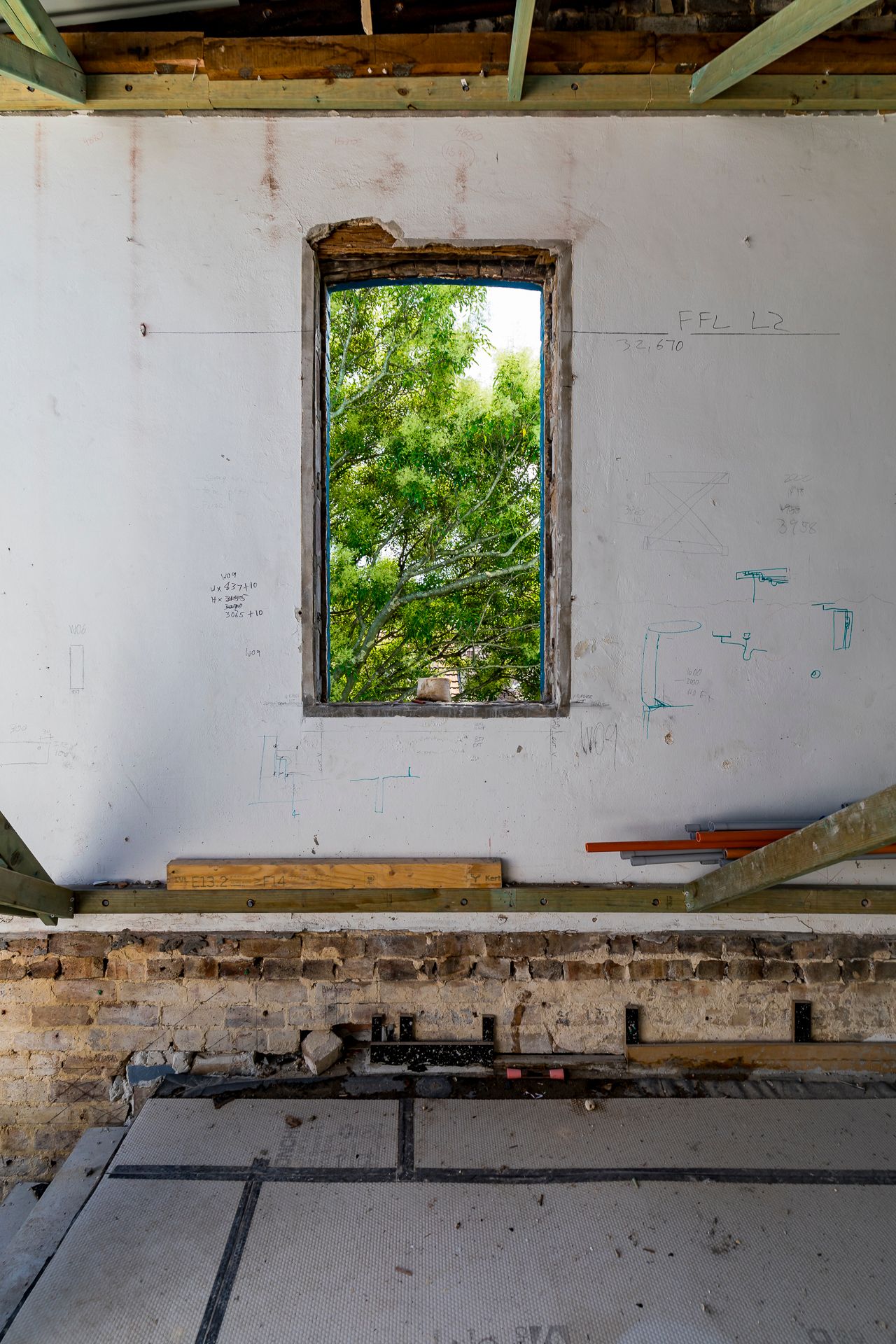 Unfinished renovation room with a bare white wall and rectangular window framing lush green trees, exposed brick and timber beams overhead.