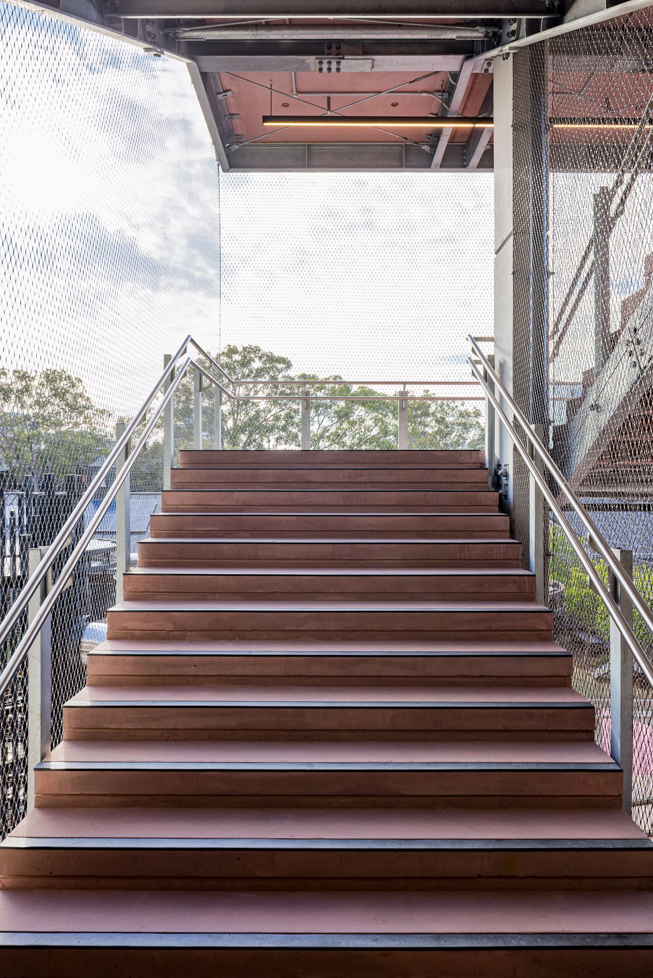 Wide timber staircase with metal handrails rising to an open landing framed by steel mesh and trees in the background.