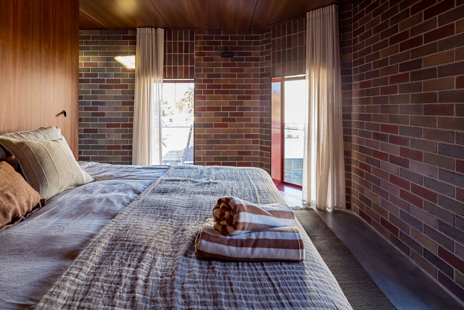 Cozy bedroom with exposed red brick walls, a large bed with gray linen bedding, rolled towels and sheer curtains beside windows.