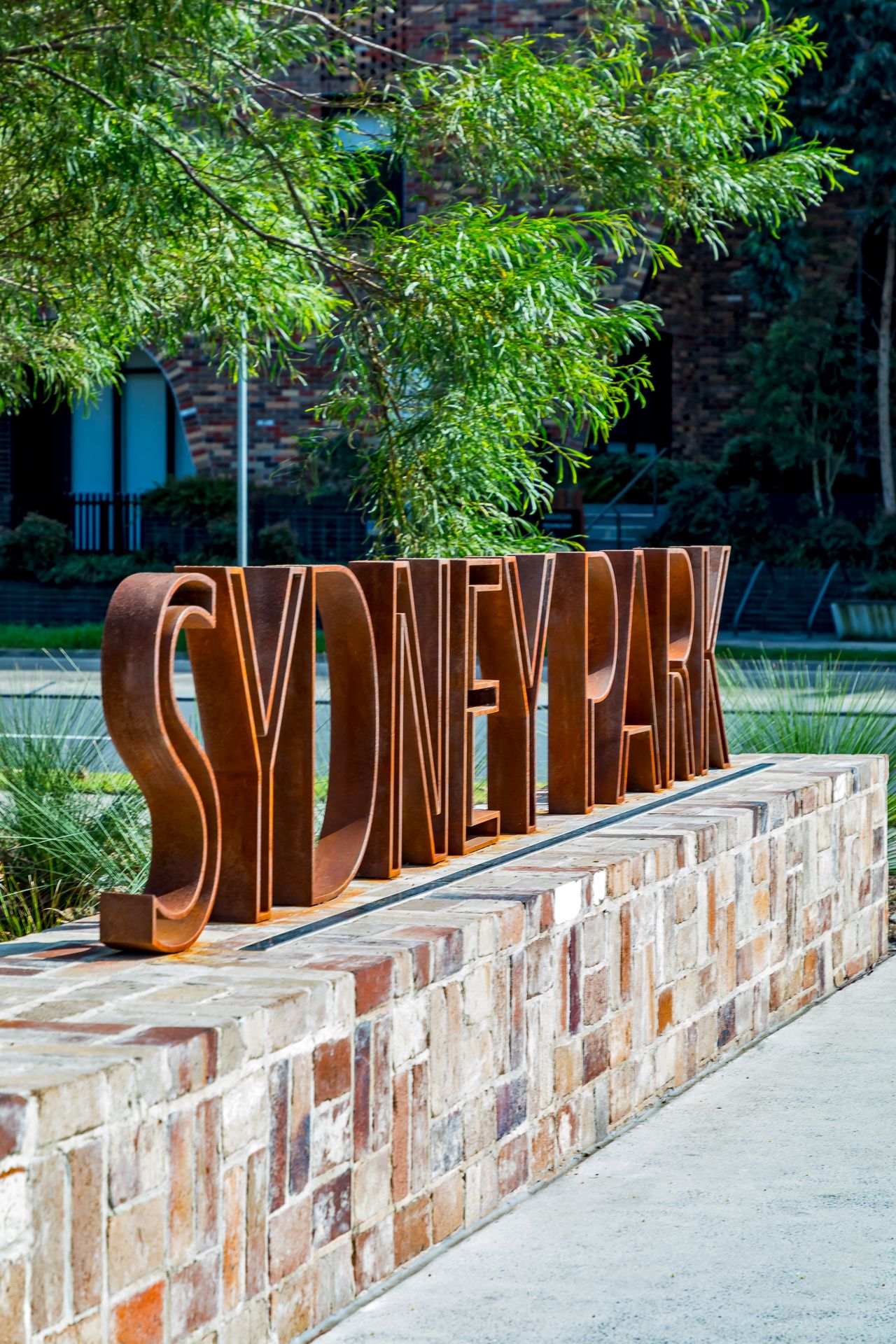 Large rusted steel letters spelling Sydney Park stand on a sandstone brick wall with trees and brick buildings behind.