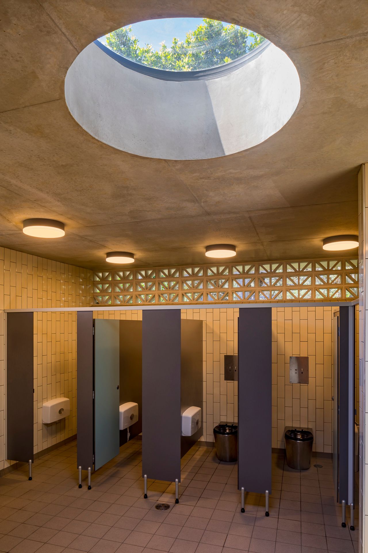 Restroom interior with a round skylight in a concrete ceiling, dark toilet cubicles, cream tiled walls and a patterned vent band.