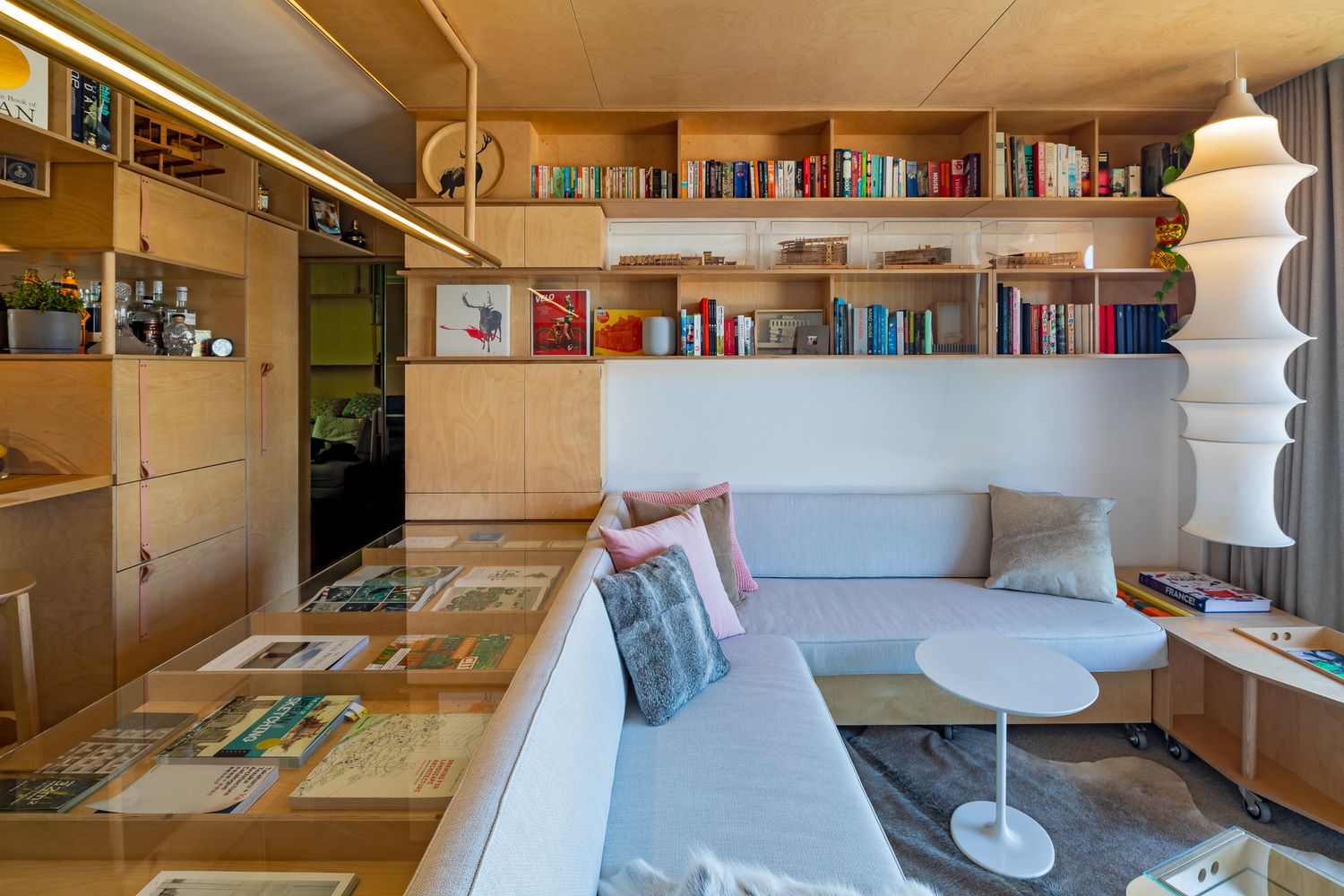 Cozy plywood-lined apartment interior with a glass-topped display table, built-in bookshelves, a gray sofa, and a white side table.