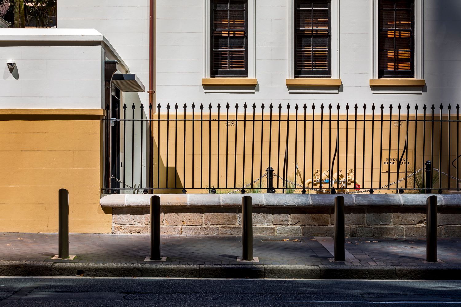 Heritage building exterior with tall windows, a black iron spiked fence, a yellow ochre wall, and bollards lining the footpath.