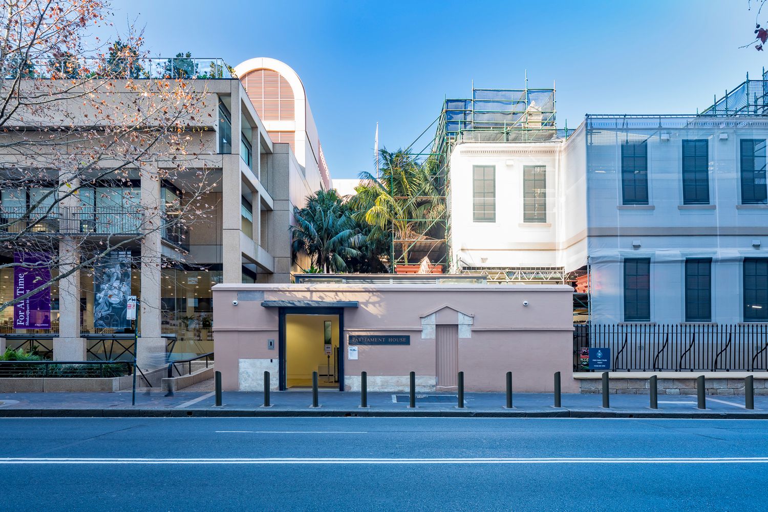 Pink modern entrance structure beside a historic white building with rooftop scaffolding, framed by palm trees and a city street.