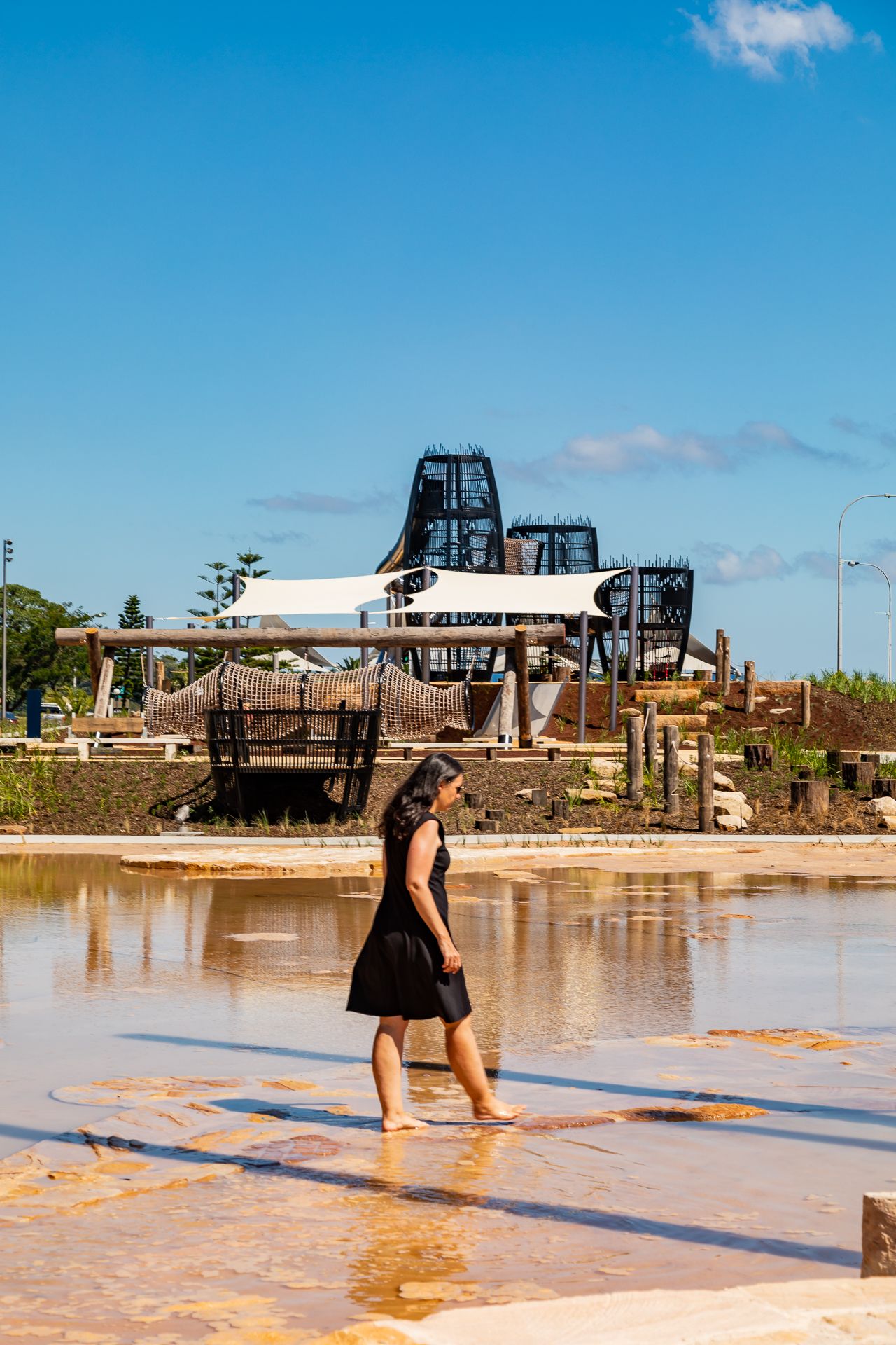 Woman in a black dress walks barefoot through shallow reflective water at a waterfront park with play structures and shade sails behind her.