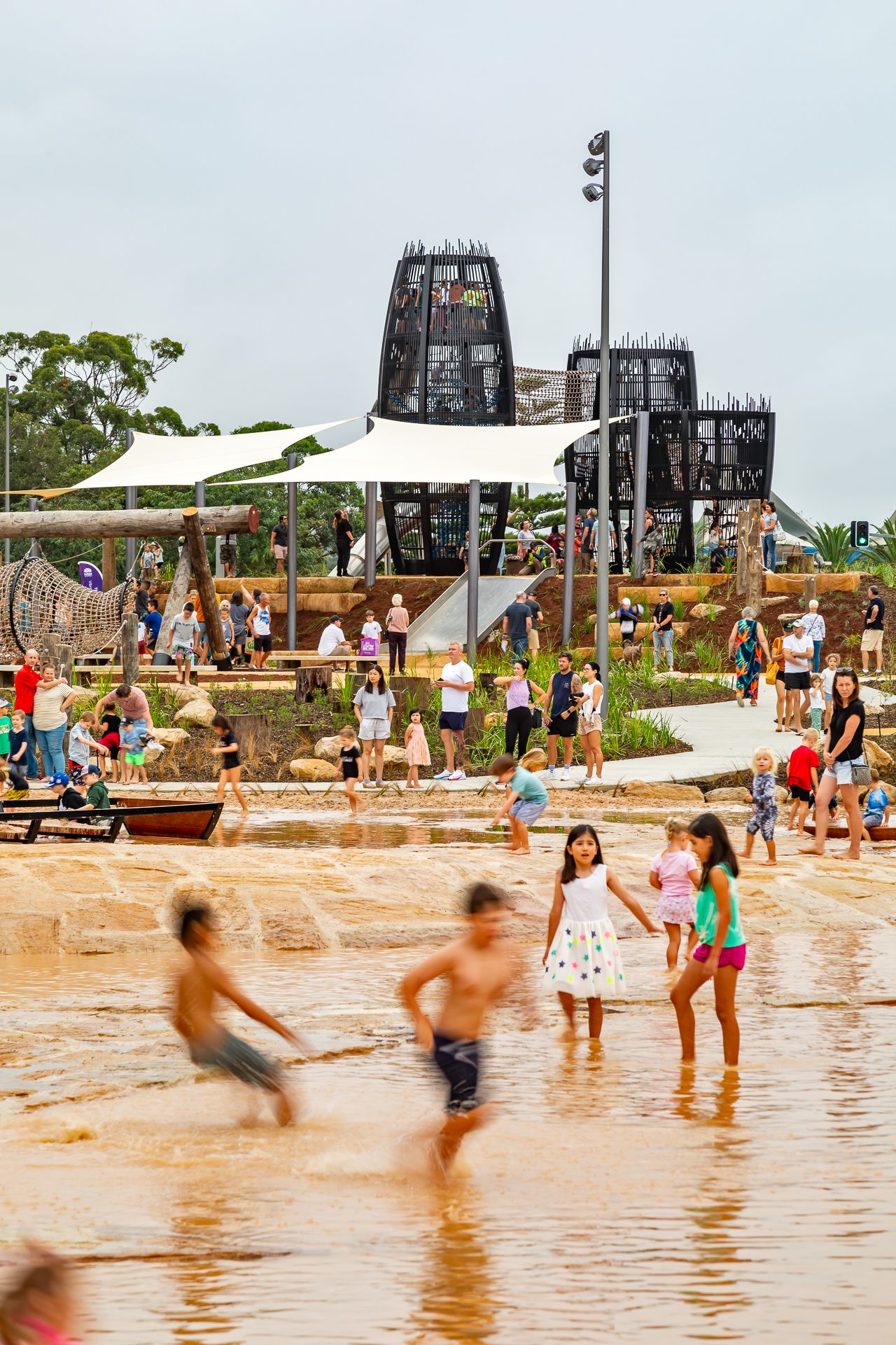 Children splash through shallow water in a public park as families watch near shade sails and dark playground towers.