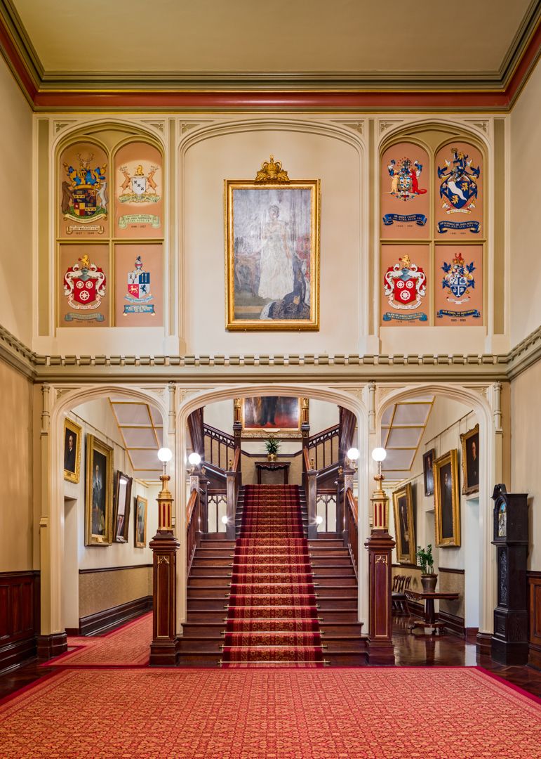 Grand heritage staircase with red carpet, framed portrait and painted coats of arms on the walls of an ornate historic interior.