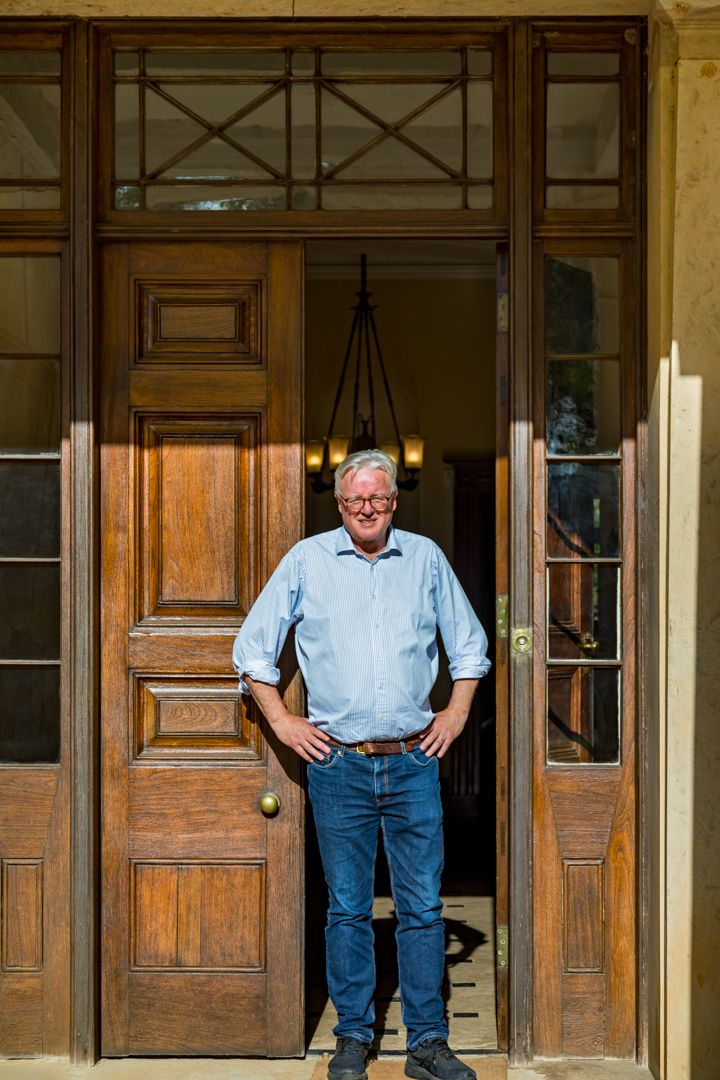 A smiling man in a light blue shirt and jeans stands with hands on hips in the doorway of a historic building with timber doors.