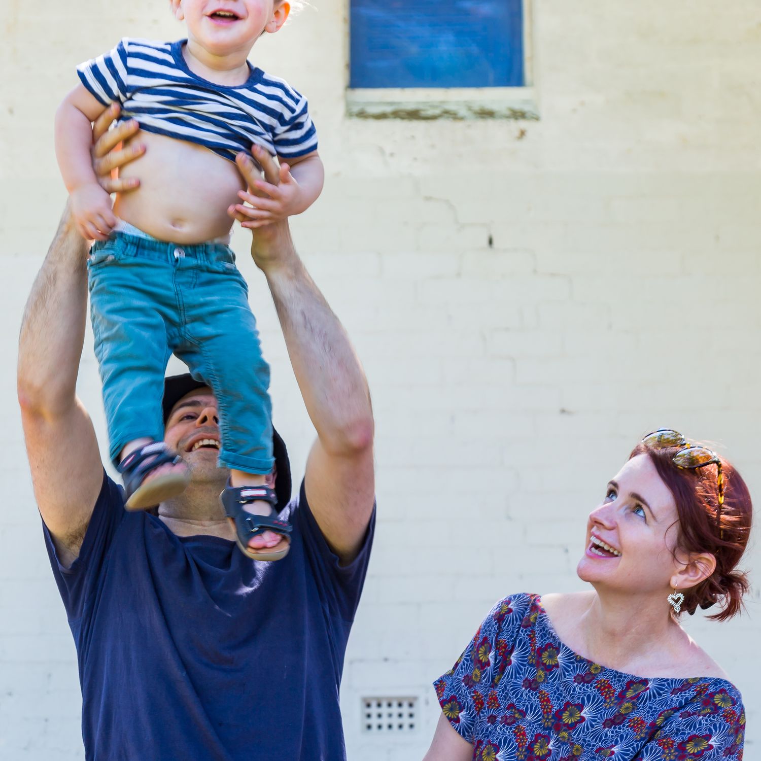 A smiling man lifts a laughing toddler in a striped shirt high into the air while a woman looks on happily against a white brick wall.