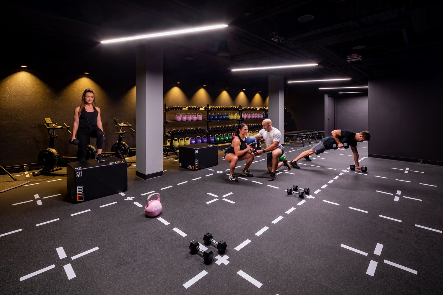 People exercise with dumbbells and bodyweight moves in a dimly lit modern gym with marked floor spots, kettlebells and weight racks.