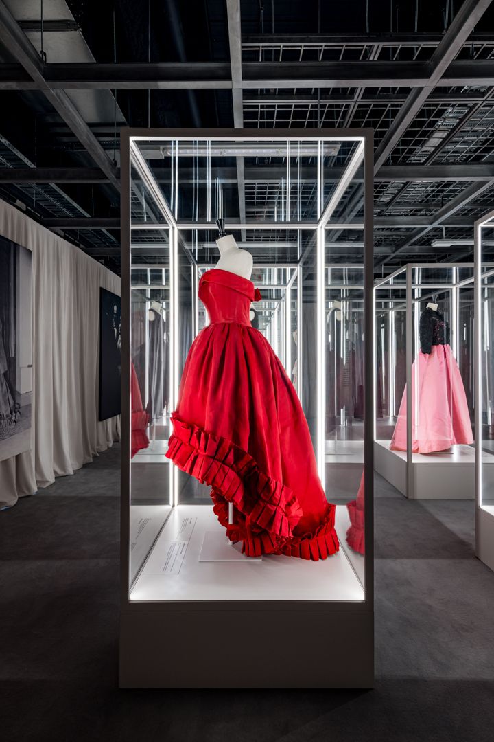 Red ruffled evening gown displayed in a lit glass case at a fashion exhibition with pink dresses in the background.
