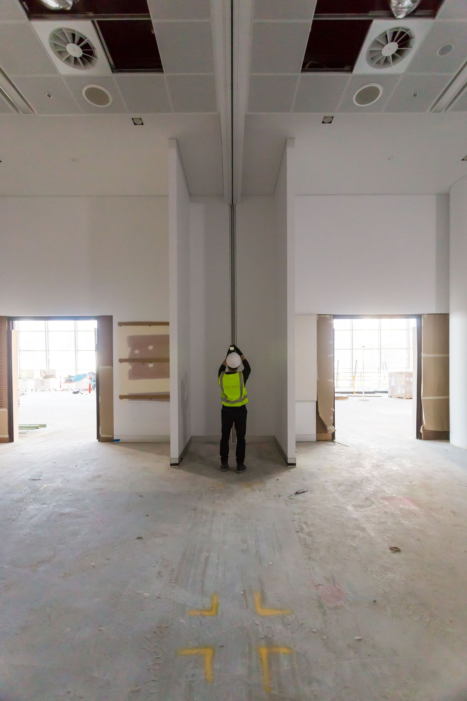 Construction worker in hi-vis vest and hard hat stands in a large unfinished hall with white plasterboard walls and open doorways.