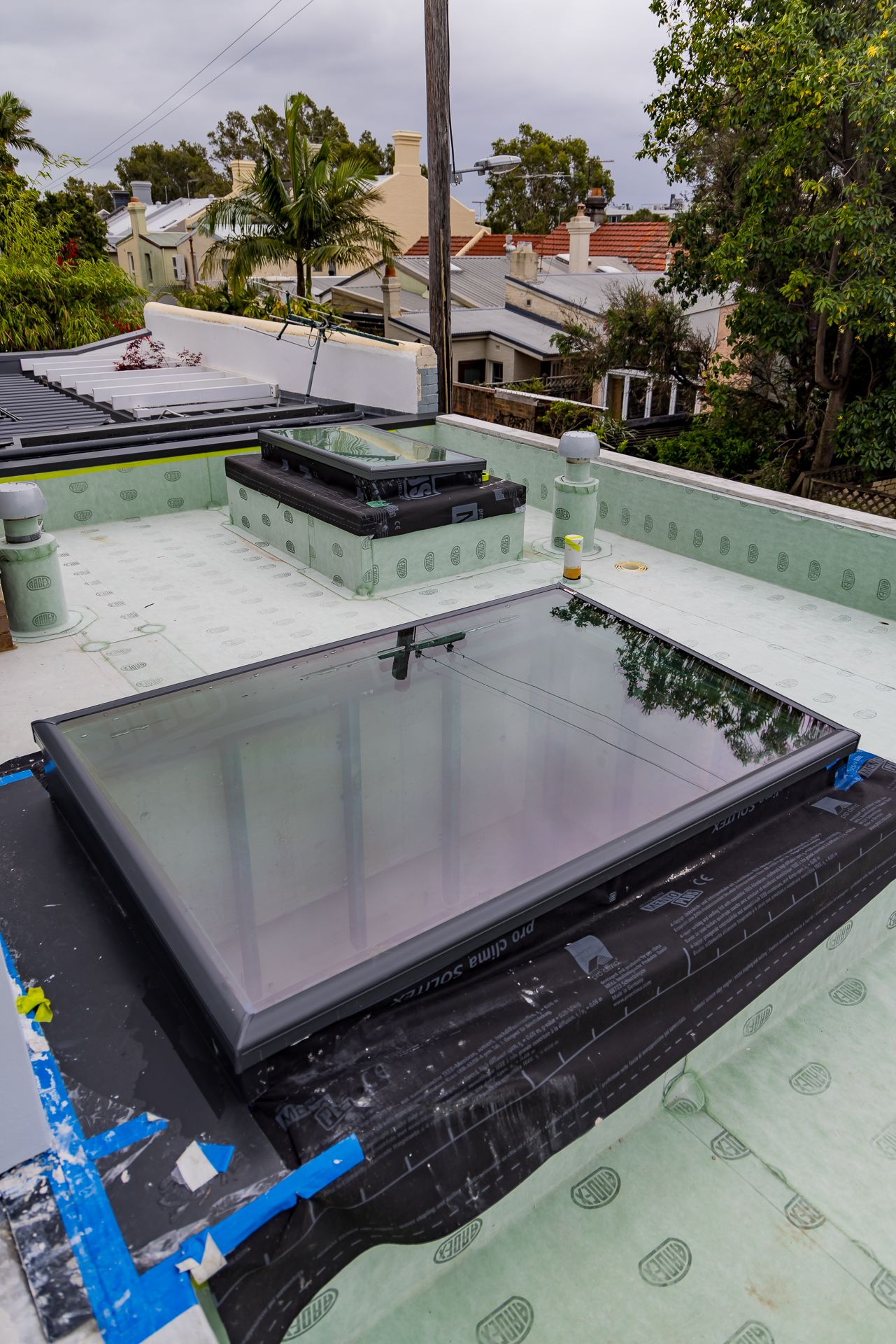 Rooftop skylight installed on a flat roof with green waterproof membrane, overlooking leafy trees and neighbouring terrace houses.