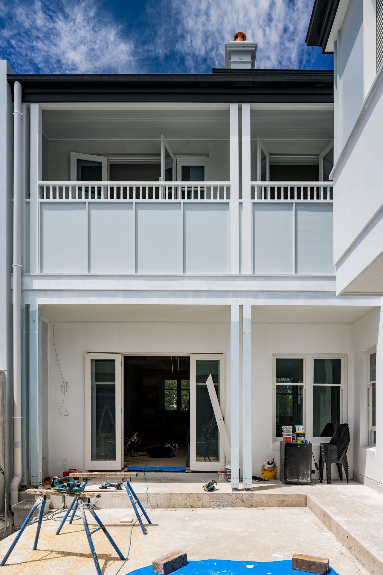 Rear of a two-storey renovated home with a white balustraded balcony, French doors and a patio area with tools and construction supplies.