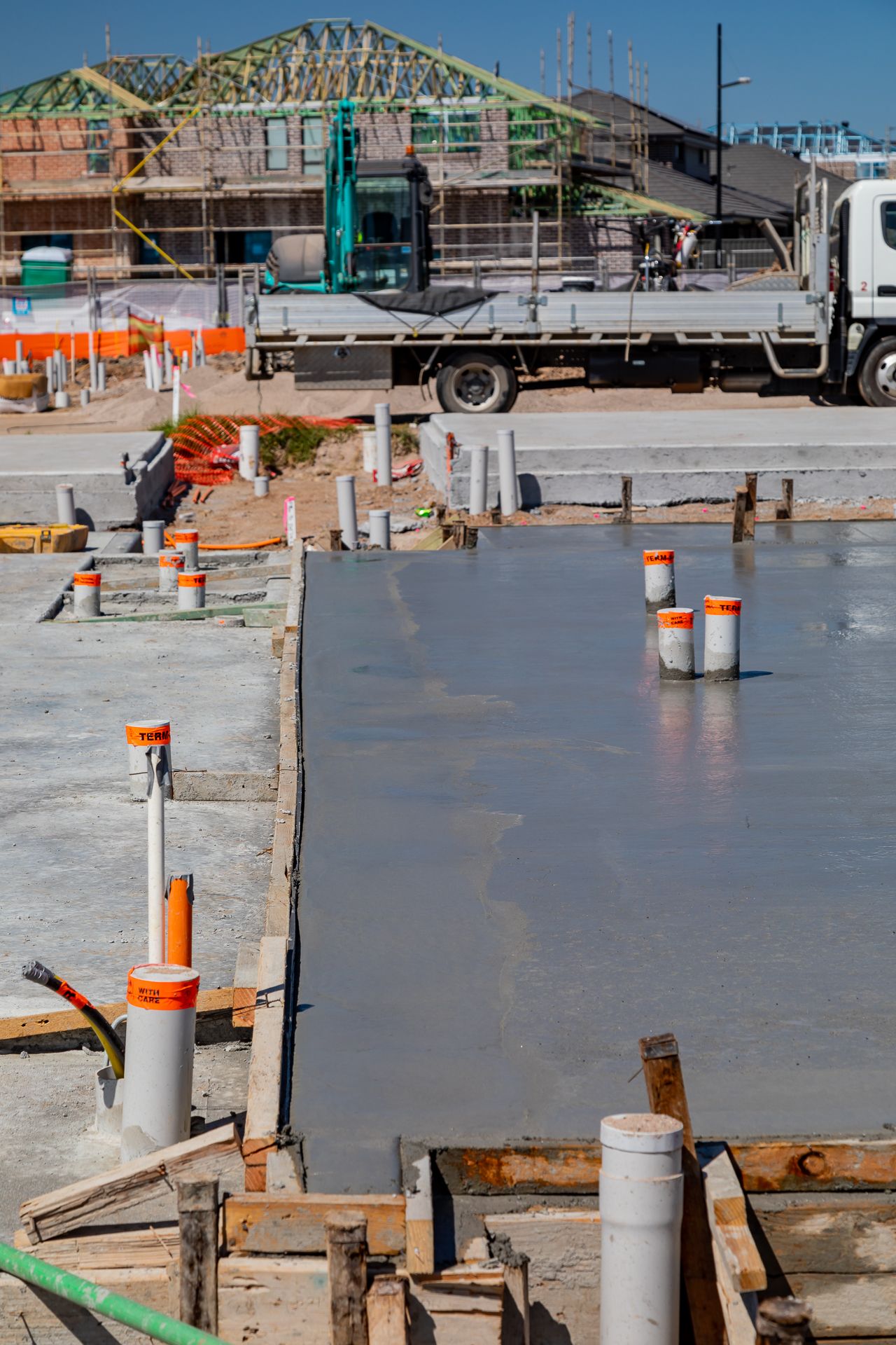 Freshly poured concrete slab with orange-capped pipes, timber formwork and a truck beside a timber-framed house under construction.