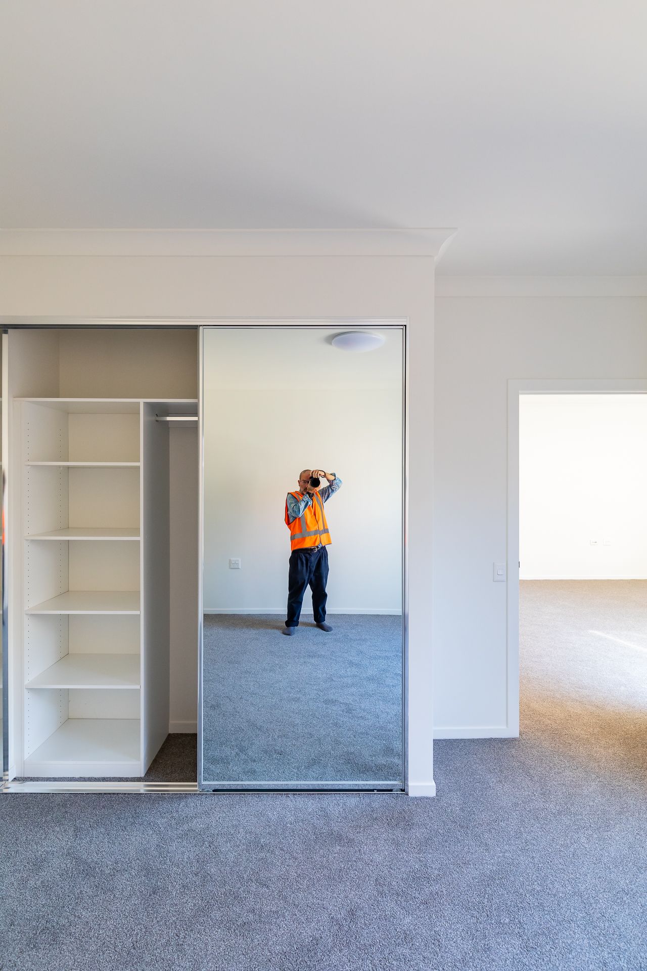 Empty bedroom with built-in wardrobe and mirrored sliding doors reflecting a worker in an orange safety vest taking a photo.