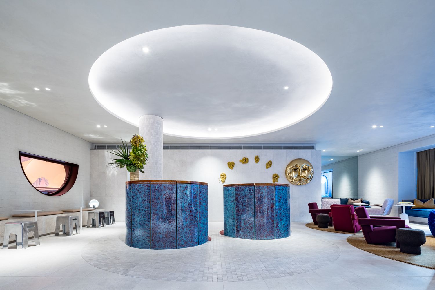 Bright hotel lobby with a glowing oval ceiling light, two blue tiled reception desks, a potted plant and colorful lounge chairs.