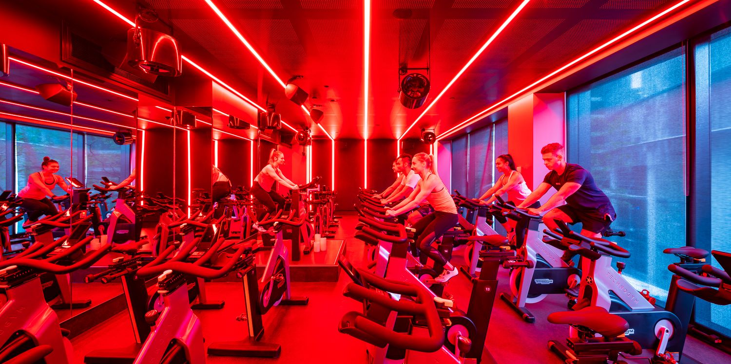 Group of riders on stationary bikes in a spin class lit by vivid red lighting, with large windows along the wall.