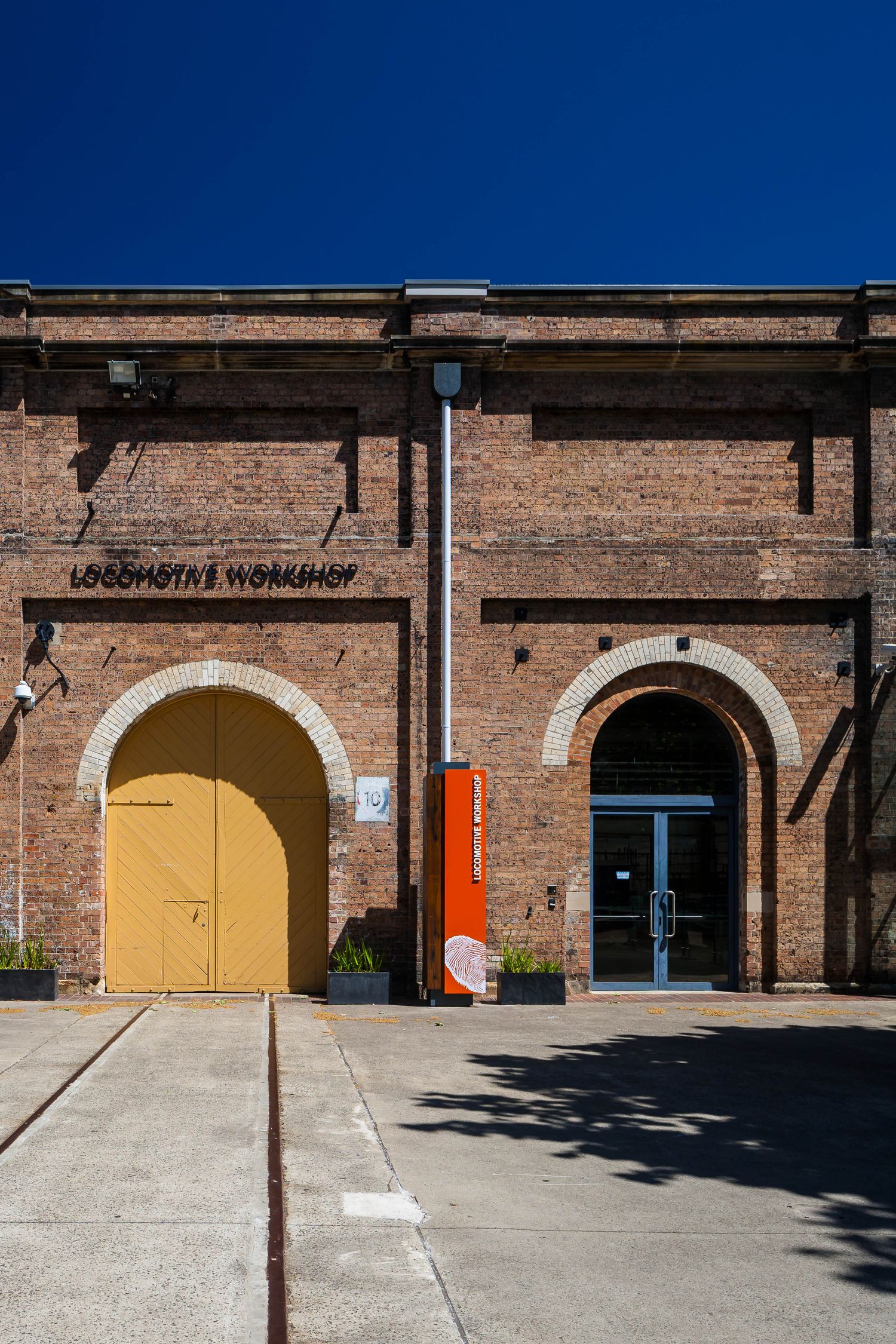 Red brick railway building with a yellow arched door, a blue glass entrance, and Locomotive Workshop lettering under a bright blue sky.