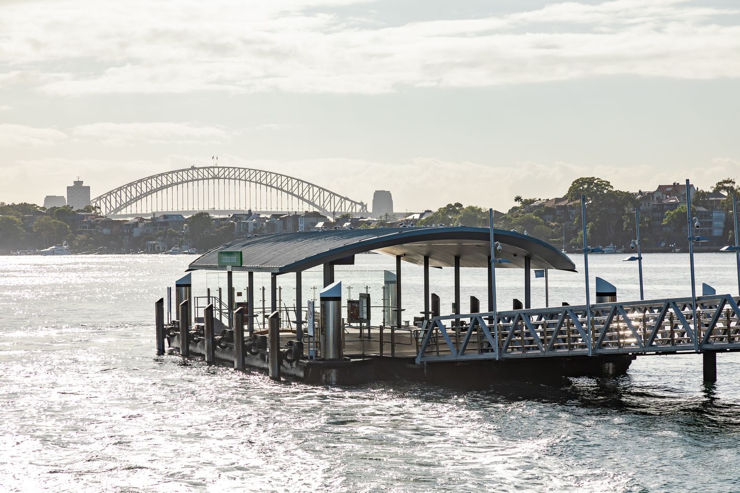 Floating ferry wharf with a curved roof on sparkling harbour water, with the Sydney Harbour Bridge visible in the distance.