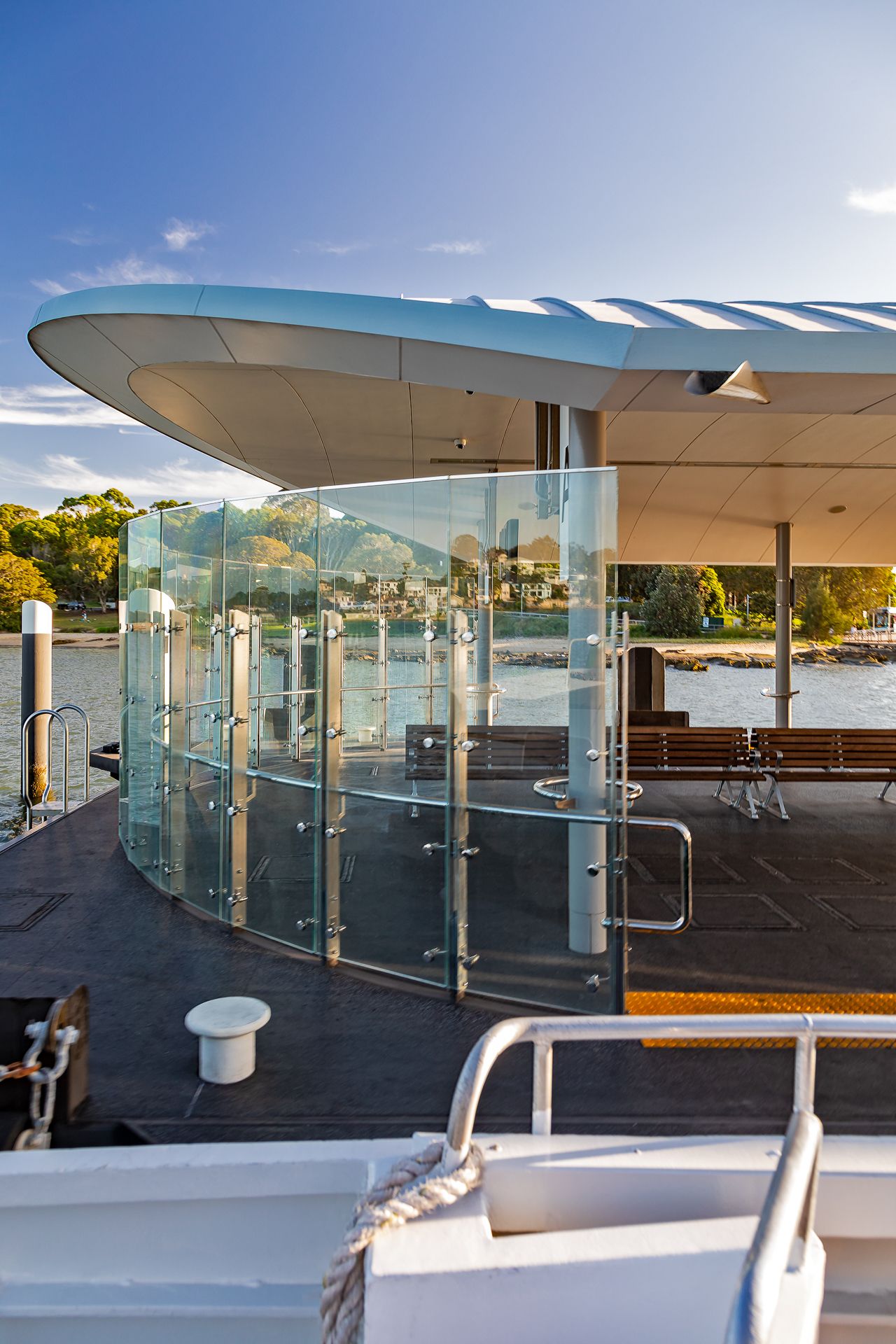 Glass-panelled waiting shelter with timber benches and a sweeping curved roof on a ferry wharf at sunset.