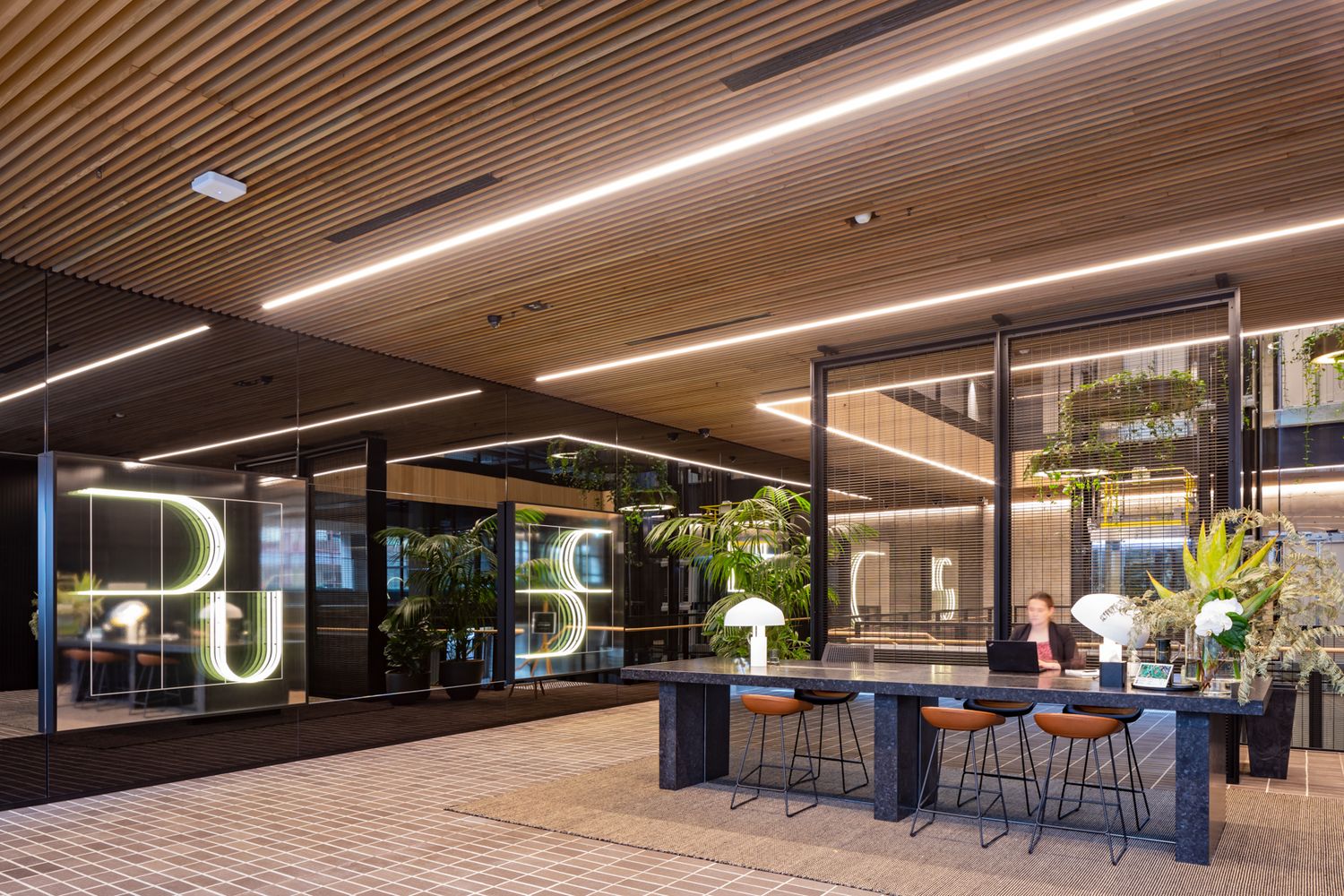 Stylish office lobby with timber slat ceiling, illuminated lettering, indoor plants and a communal high table with stools.