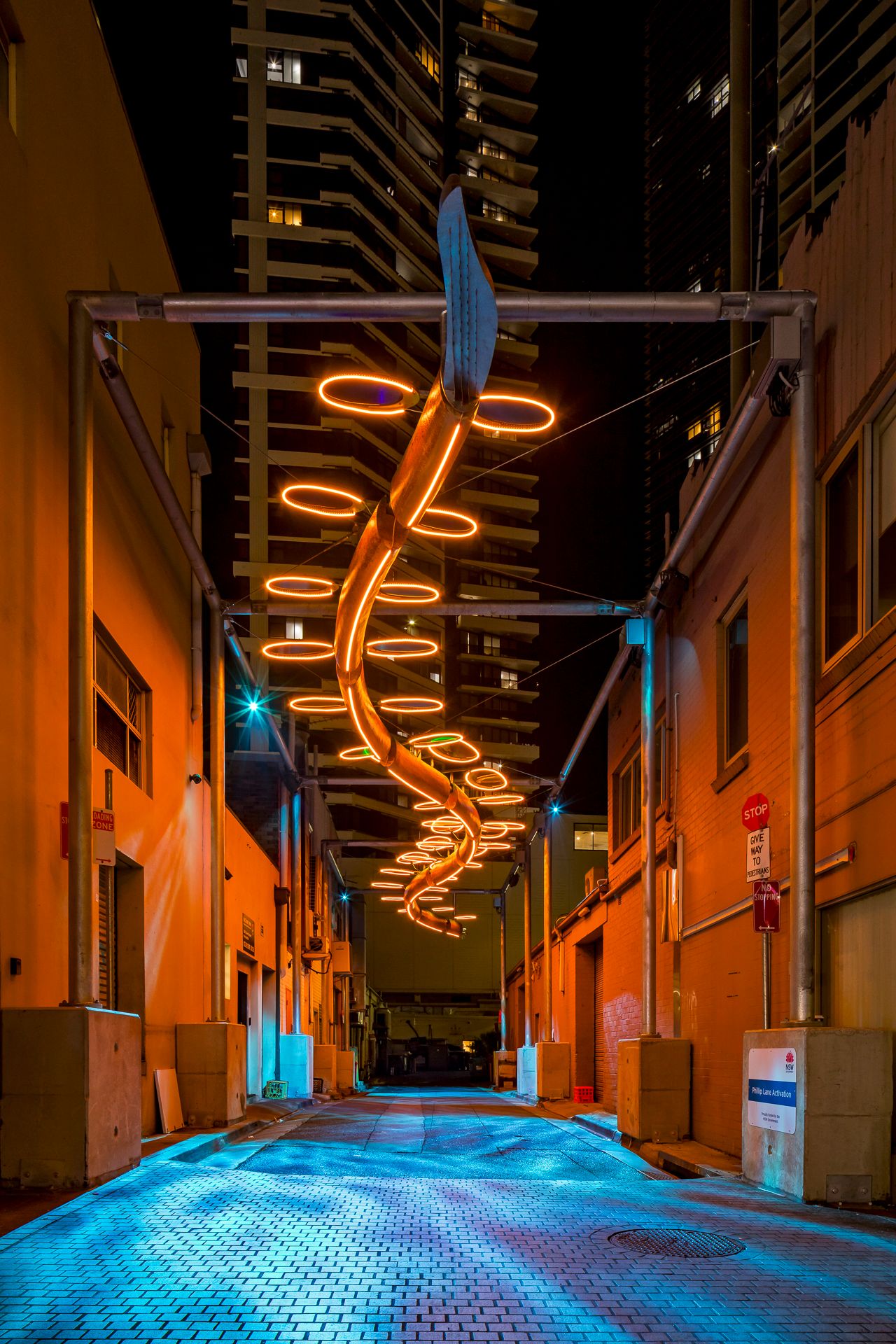 An orange neon spiral sculpture hangs over a paved alley, casting warm light on the walls beside tall towers.