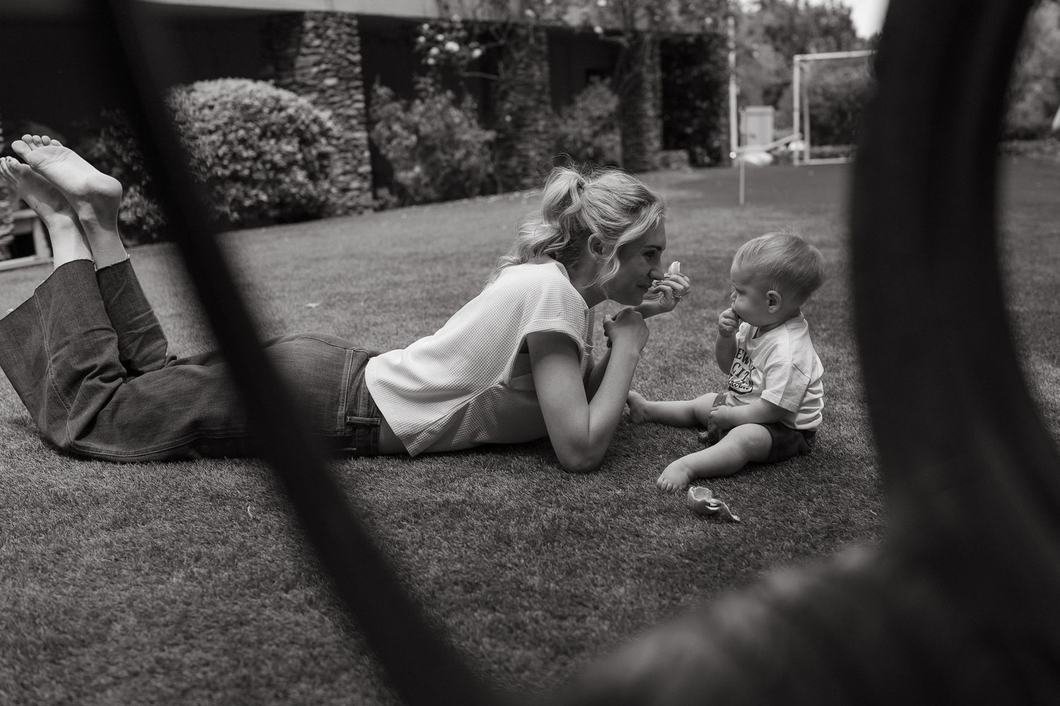 Black and white photo of a mother lying on the grass playing with her toddler, framed by a dark circular edge.