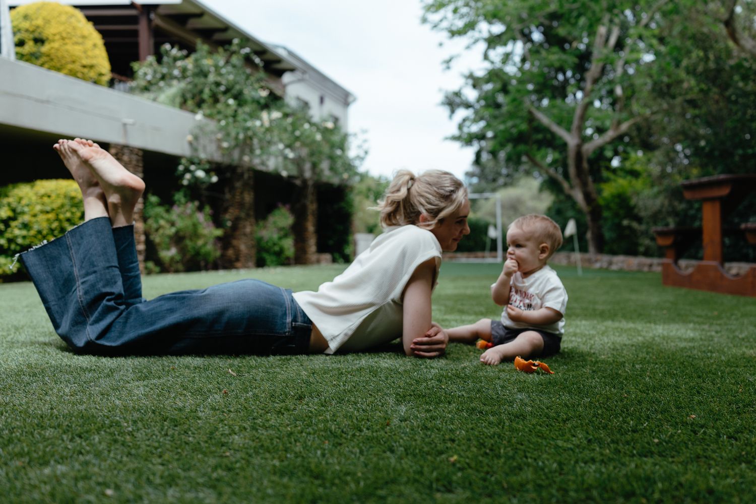 Mother lies on a green lawn while her baby sits beside her, with trees and a house in the background.