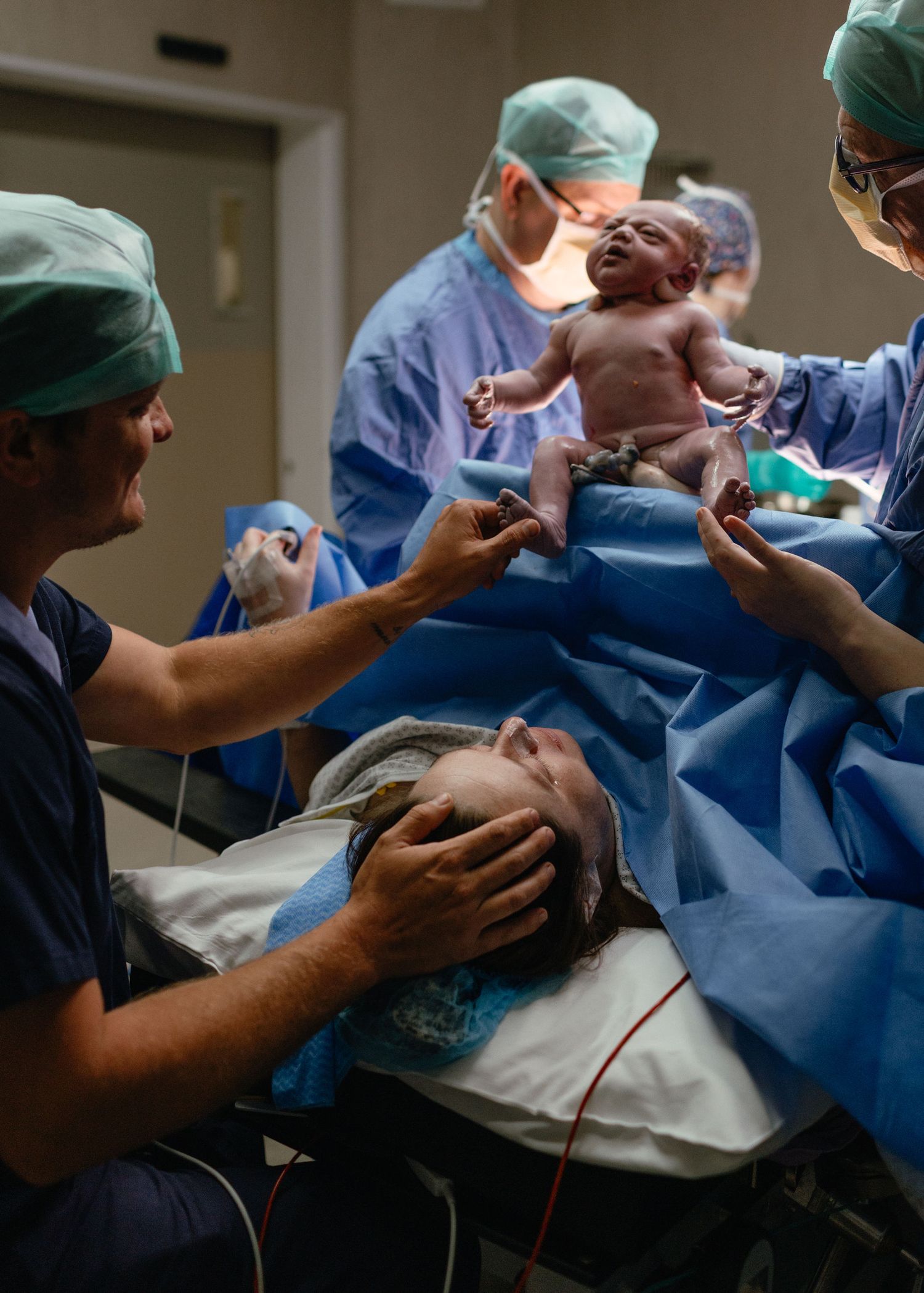 Surgeons in scrubs lift a newborn baby over a blue surgical drape while the mother lies on the operating table during a cesarean birth.