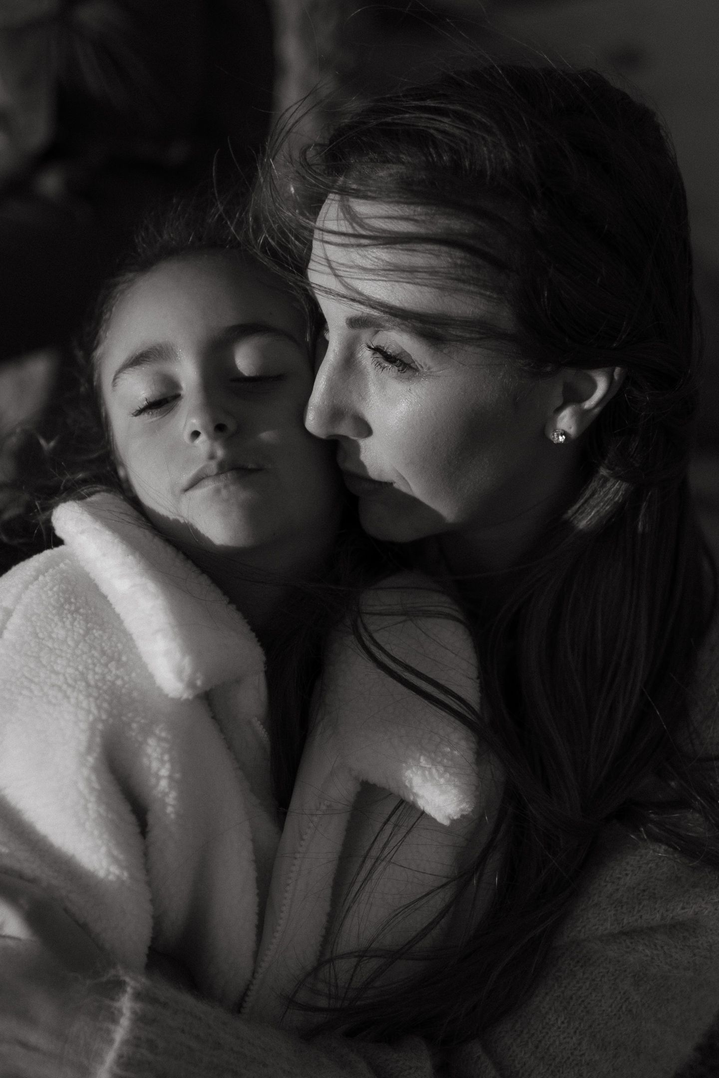 Black and white portrait of a mother tenderly holding her sleeping daughter in a soft fleece jacket.