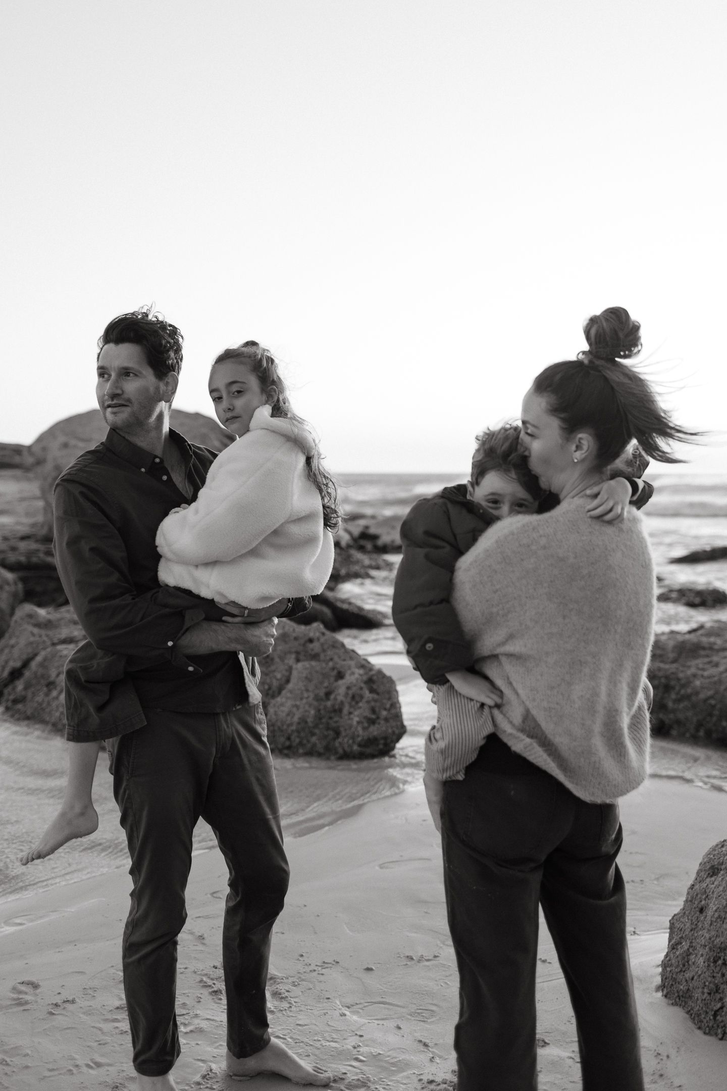 Black and white photo of a family on a rocky beach, parents holding their two children close at dusk.