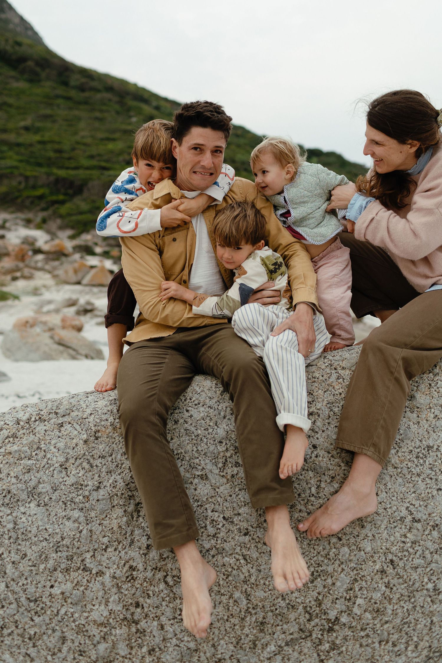 A laughing father is hugged by his children and wife as the family cuddles on a granite rock by the sea.