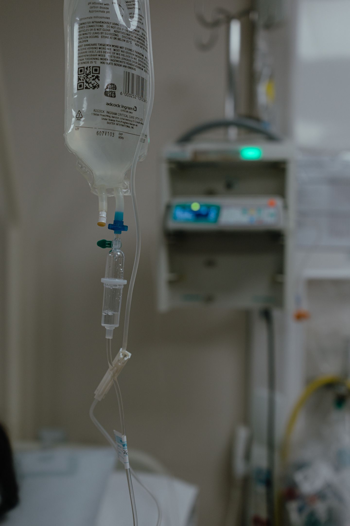 Hanging IV fluid bag with drip chamber and tubing in a hospital room, with a blurred infusion pump glowing green in the background.