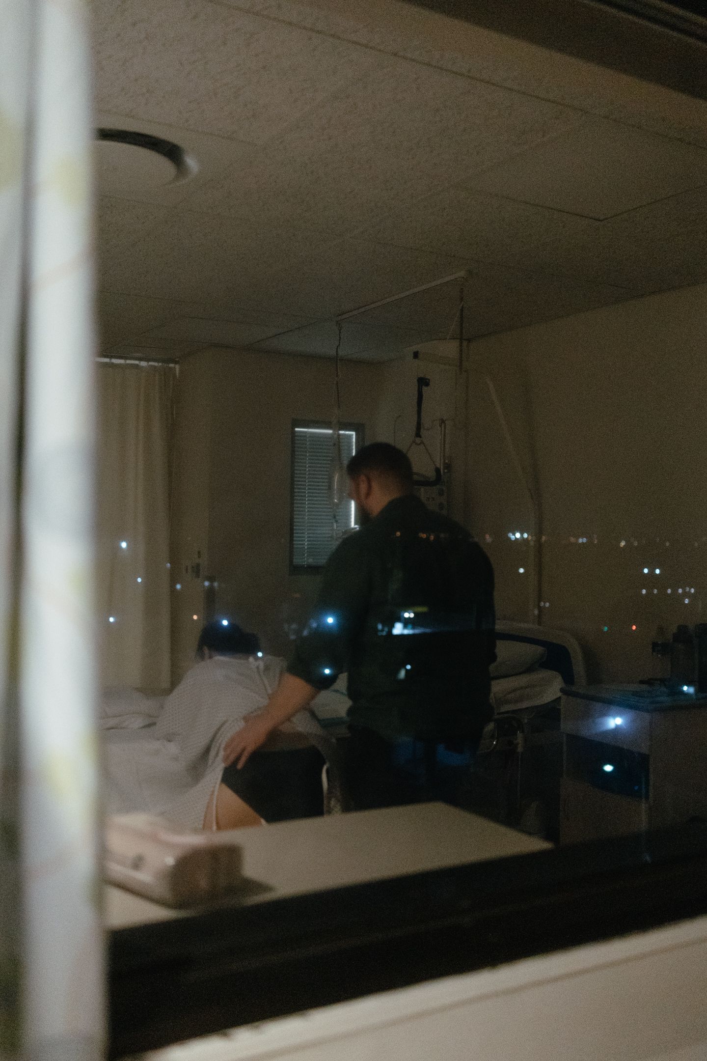 Dimly lit hospital room at night, seen through a window, where a person in dark clothing tends to a patient lying in bed.
