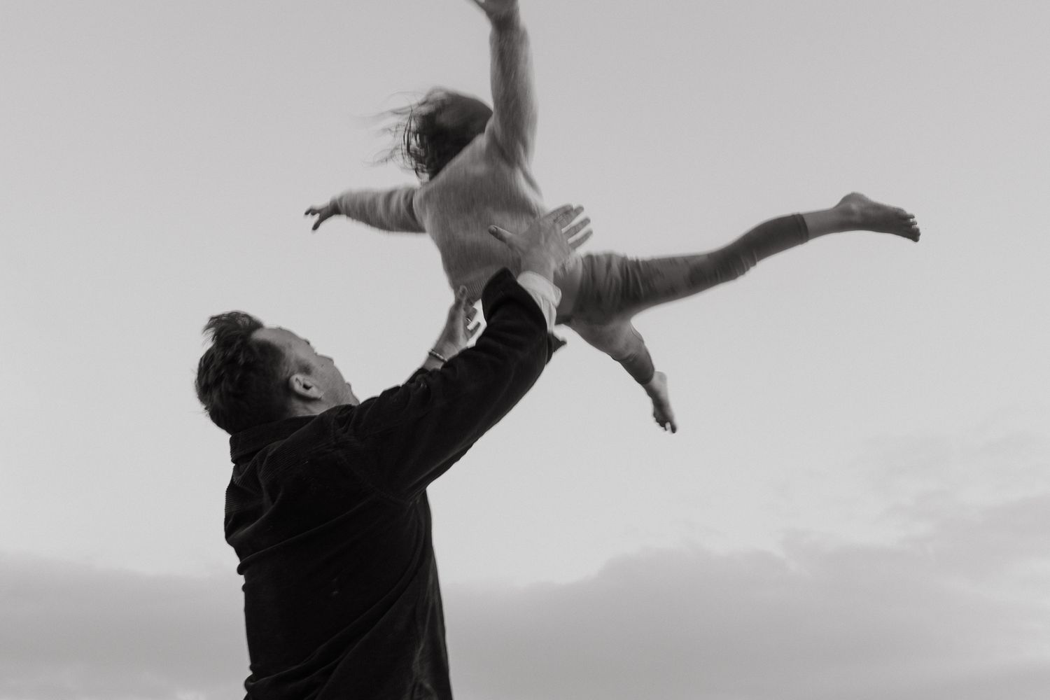 In a black and white photo, a father in a dark jacket lifts a laughing child high into the air against a cloudy sky.