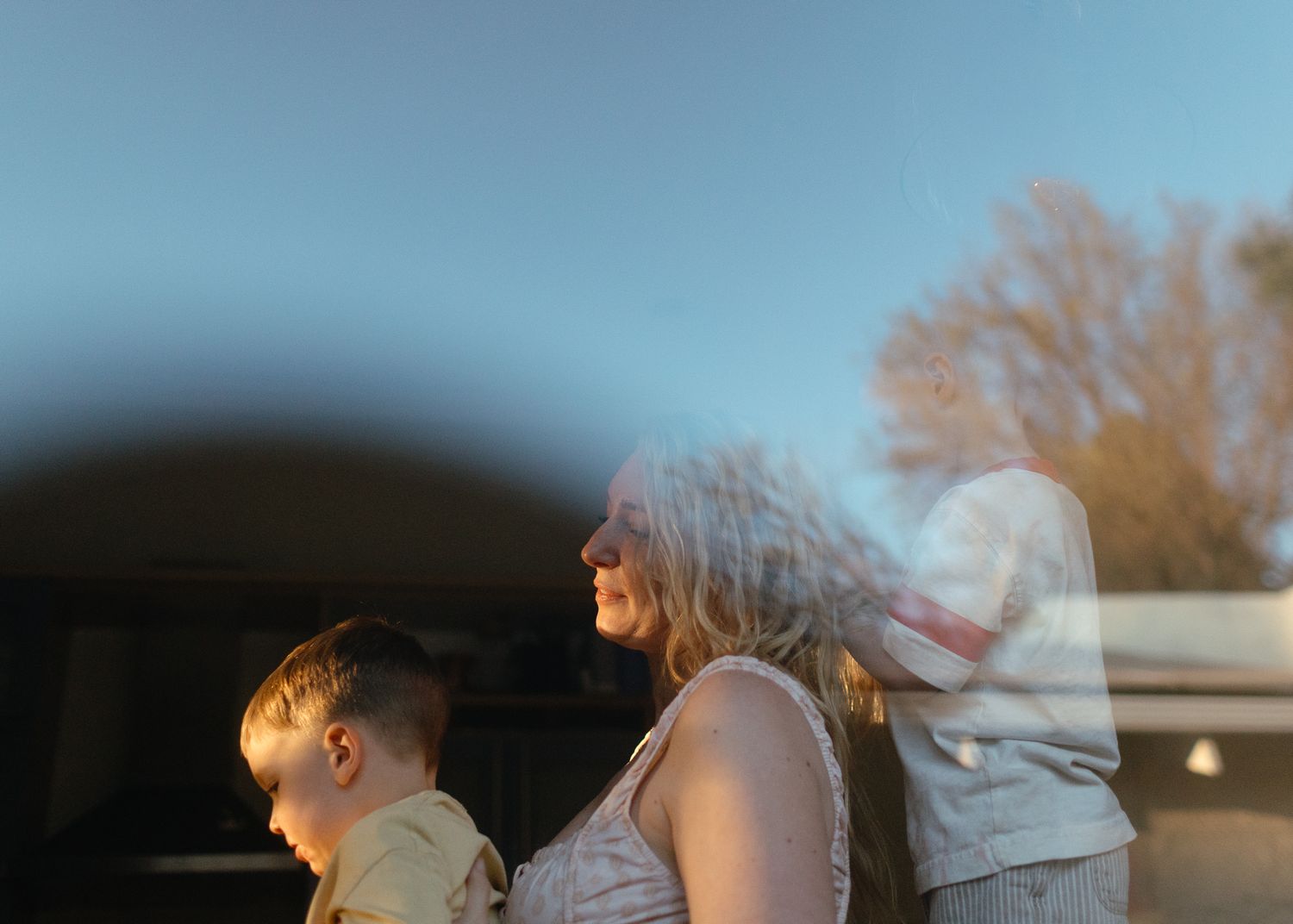 Soft reflection on glass shows a woman in profile beside a young boy, with bare trees and blue sky in the background.