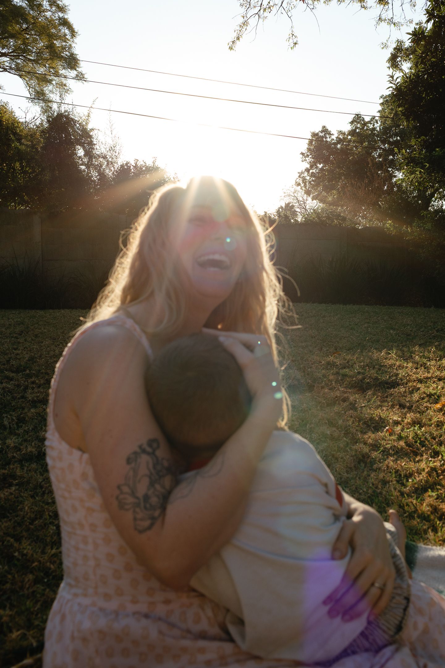 A laughing woman with a tattooed arm hugs a child in a sunlit grassy yard with golden backlight.