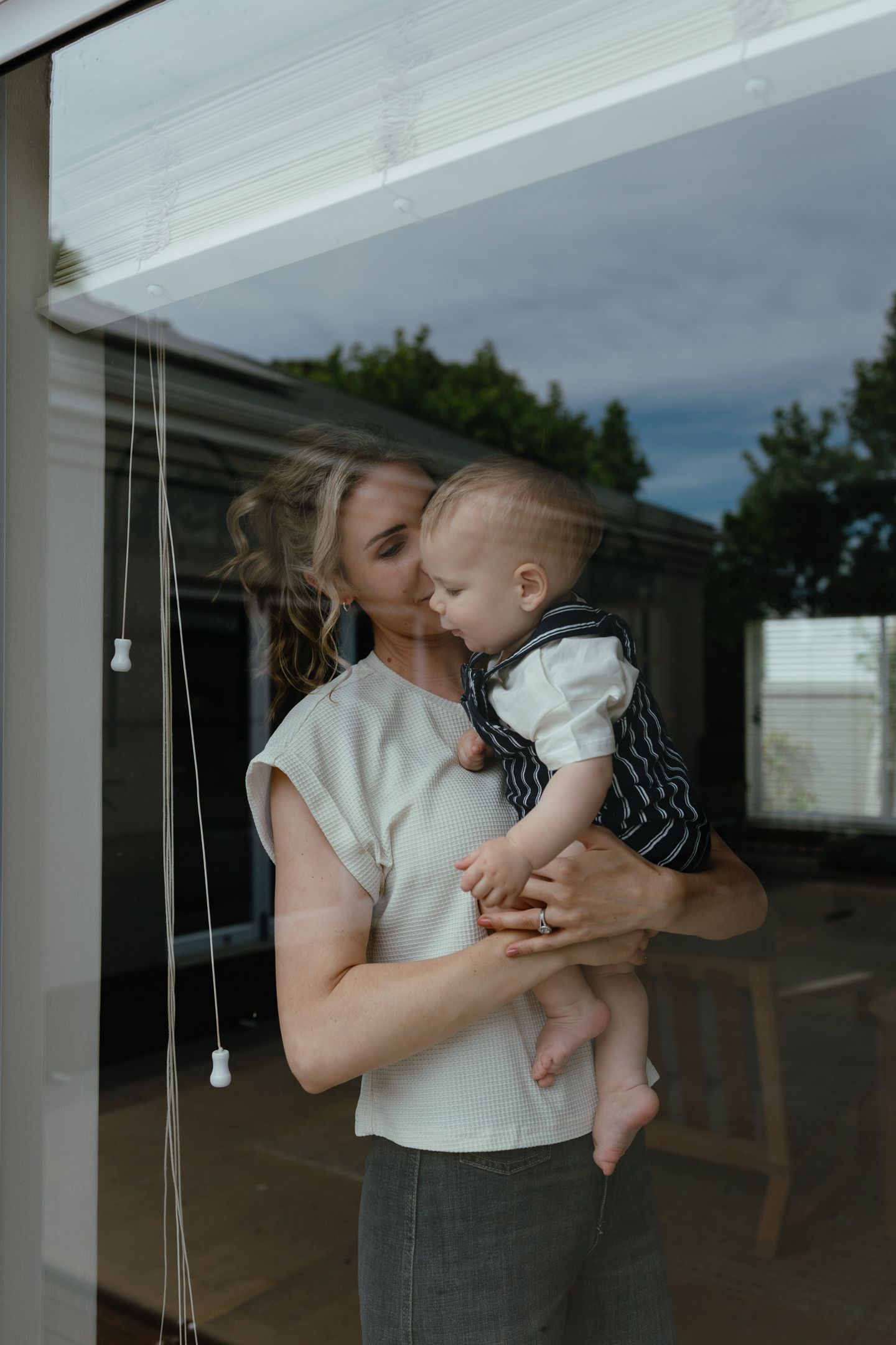 A blonde woman kisses her baby's head as they stand behind a window with soft reflections of trees and sky.
