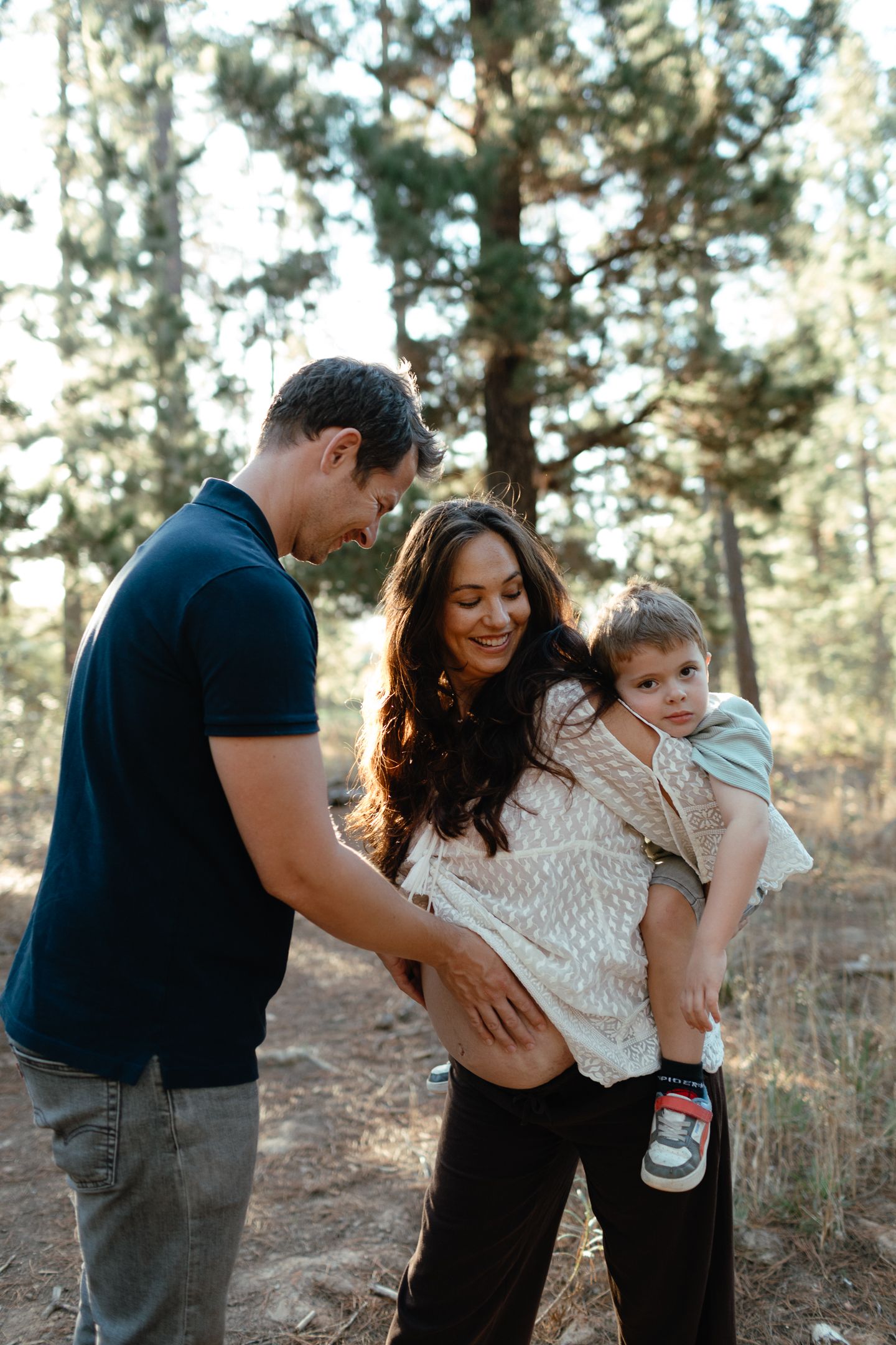 Smiling pregnant woman carries her toddler son as her husband embraces her belly in a sunlit pine forest.