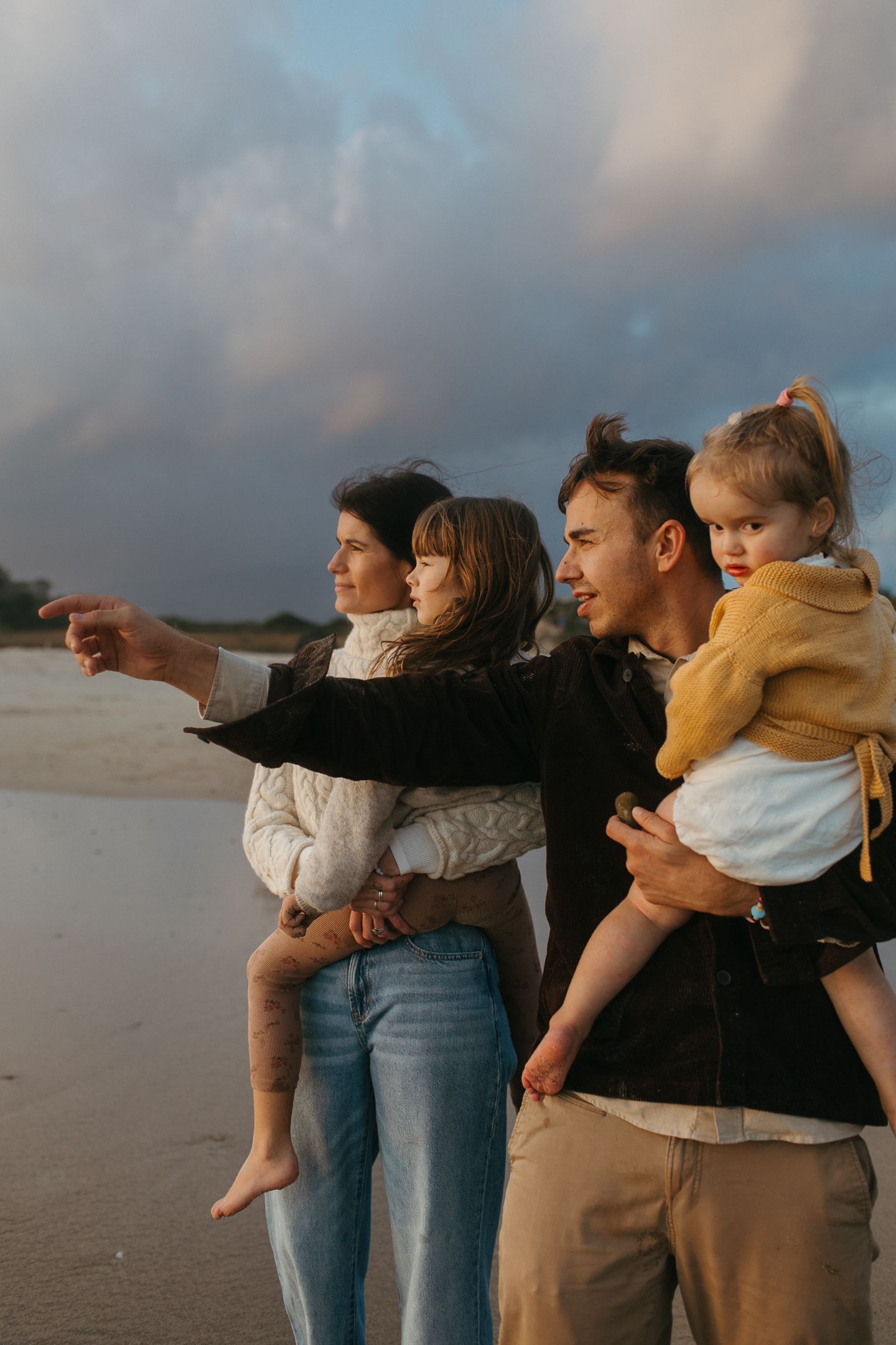A mother points toward the horizon while a father and two young daughters watch the beach sunset under dramatic clouds.