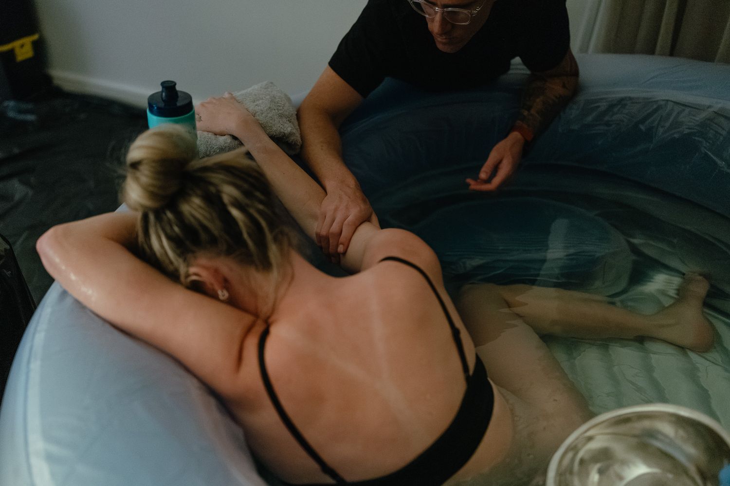 Partner massages the arm of a laboring woman resting on the edge of an inflatable birth pool during a home water birth.