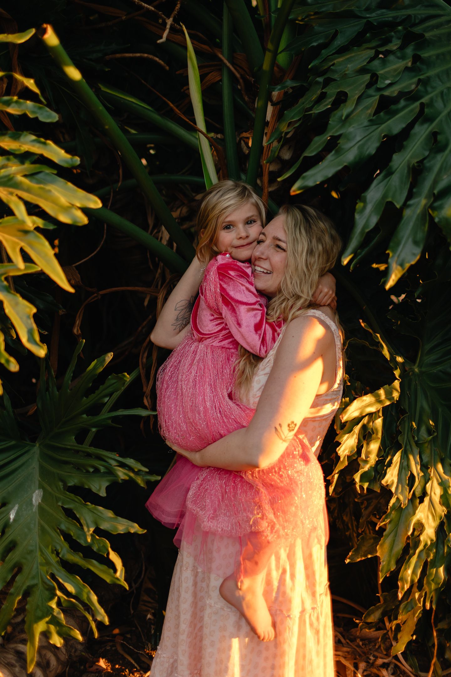 Smiling blonde woman in a light dress holds a little girl in a pink outfit among lush green tropical leaves in golden sunlight.