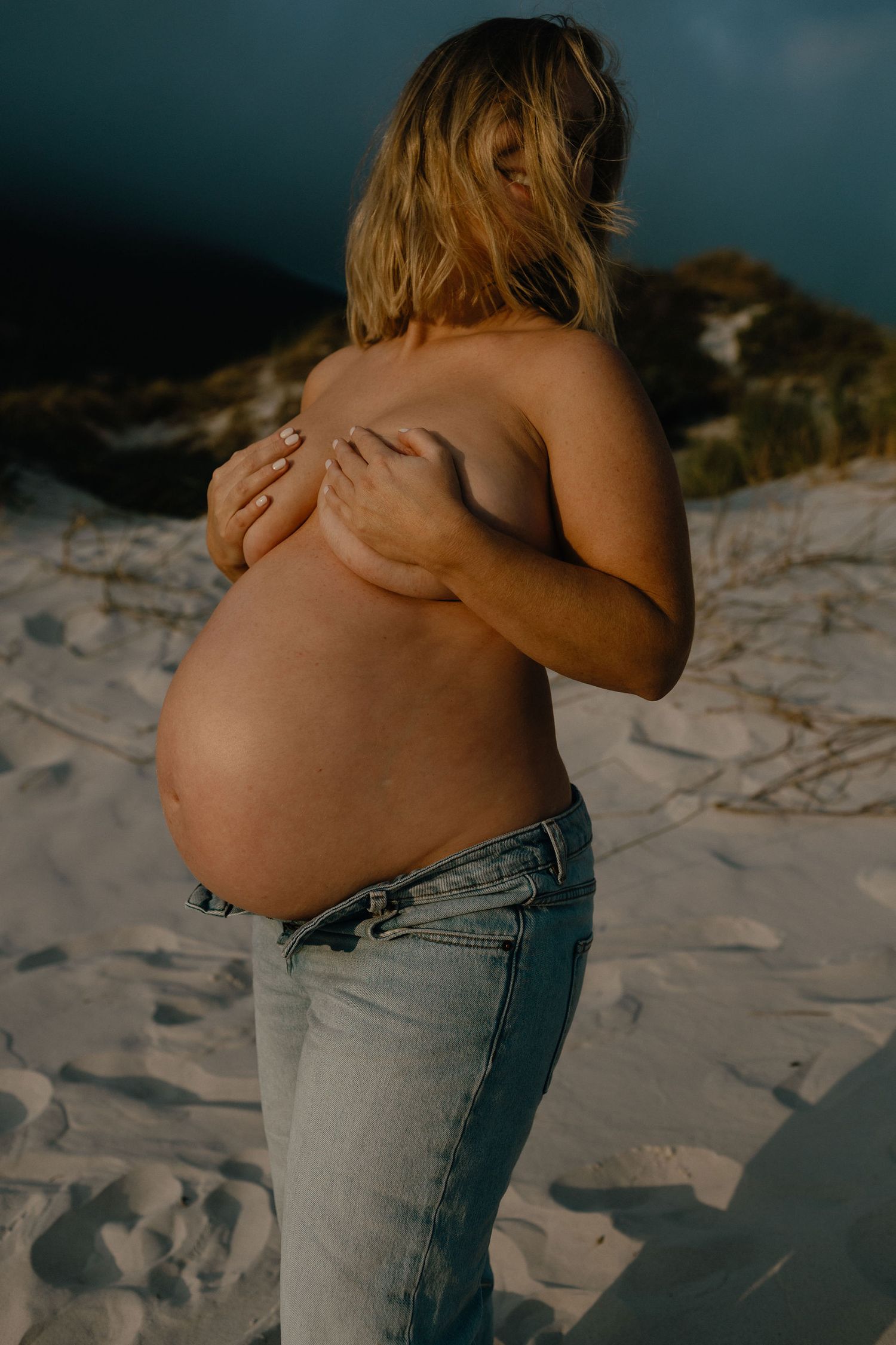 Pregnant woman in light jeans stands on a sandy beach at dusk, gently covering her chest with her hands and showing her round belly.