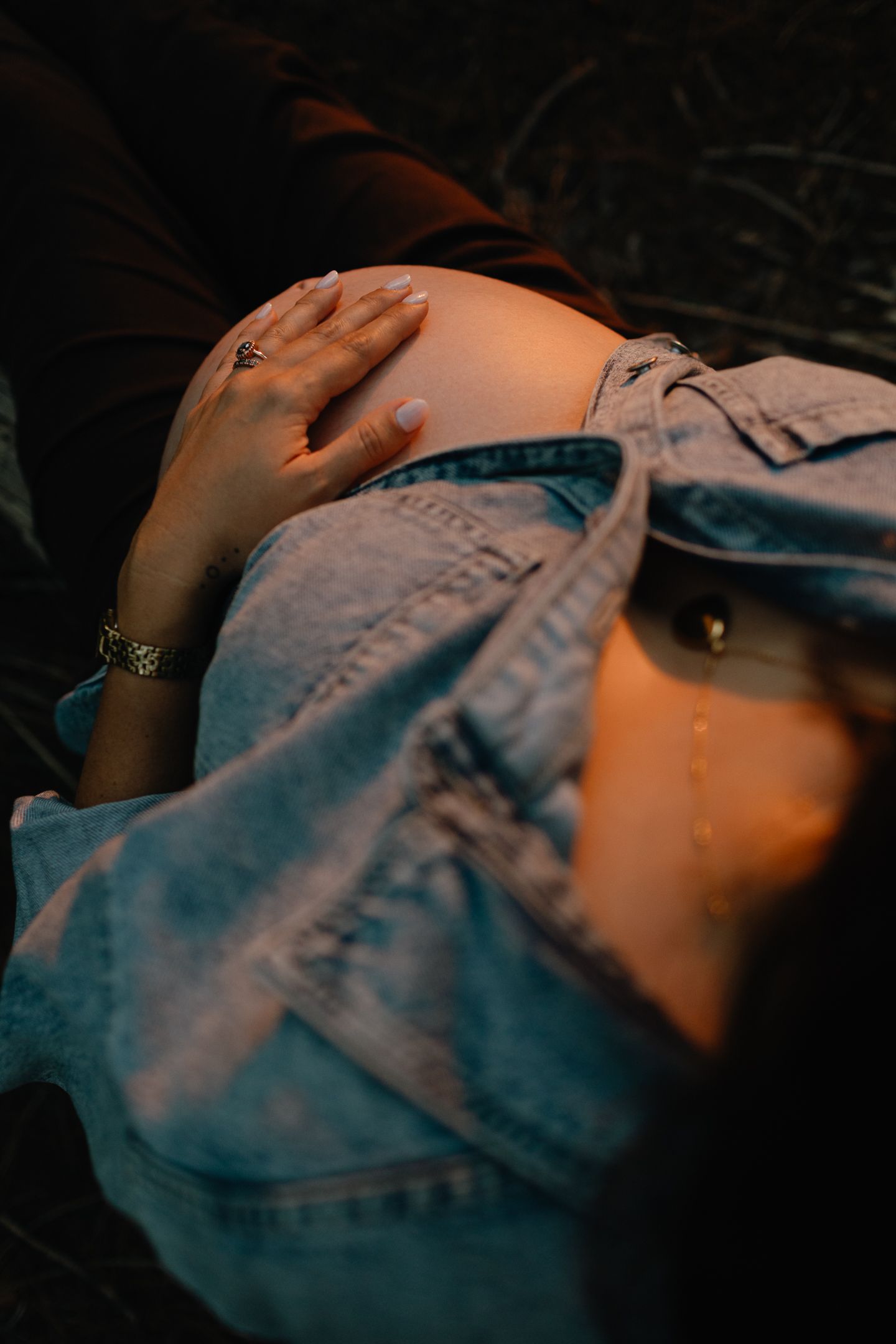 A pregnant woman in a denim jacket rests her hand on her bare belly in moody, warm evening light.