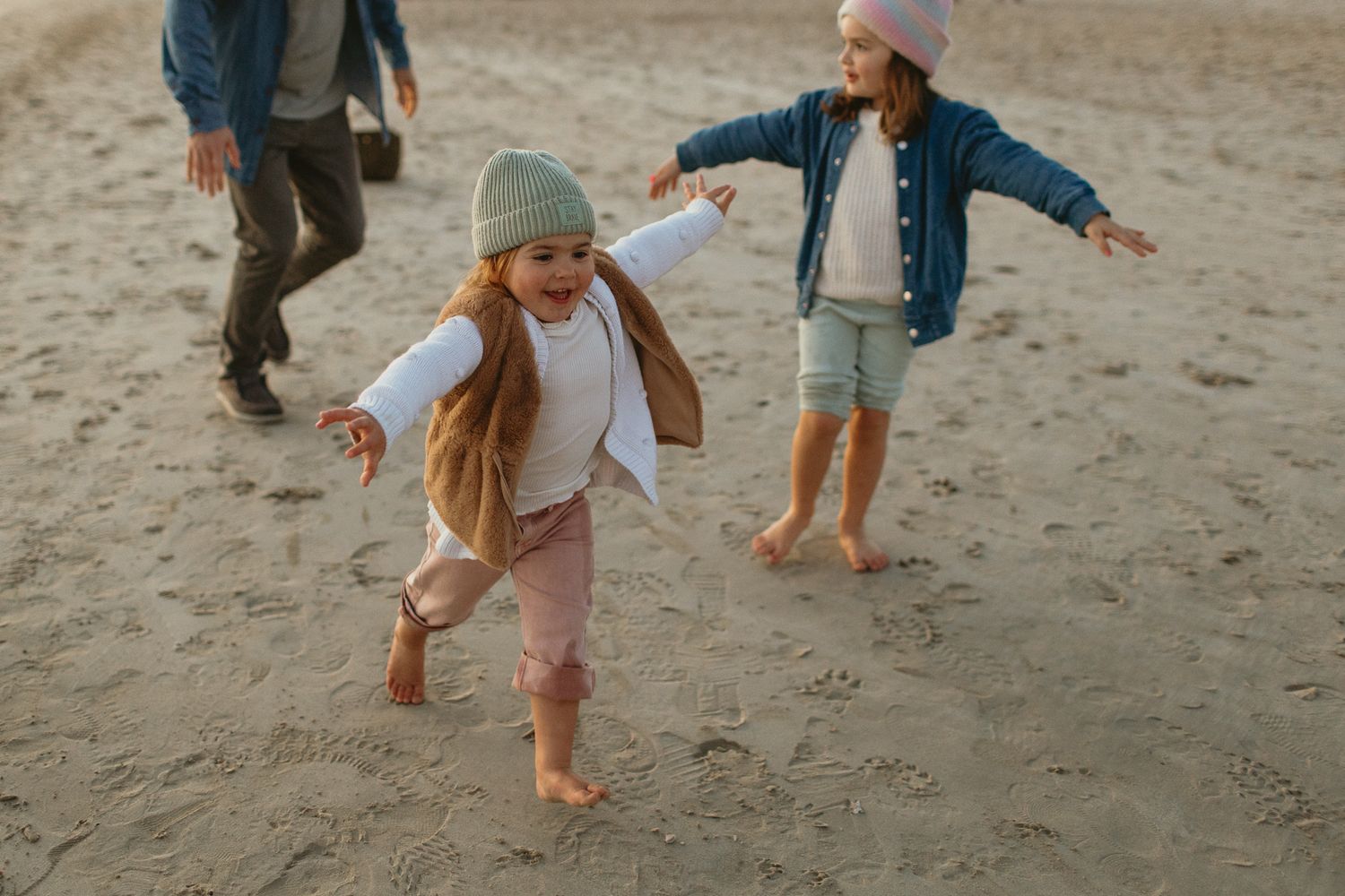 Two children in cozy beanies run and play barefoot on a sandy beach while an adult walks nearby.