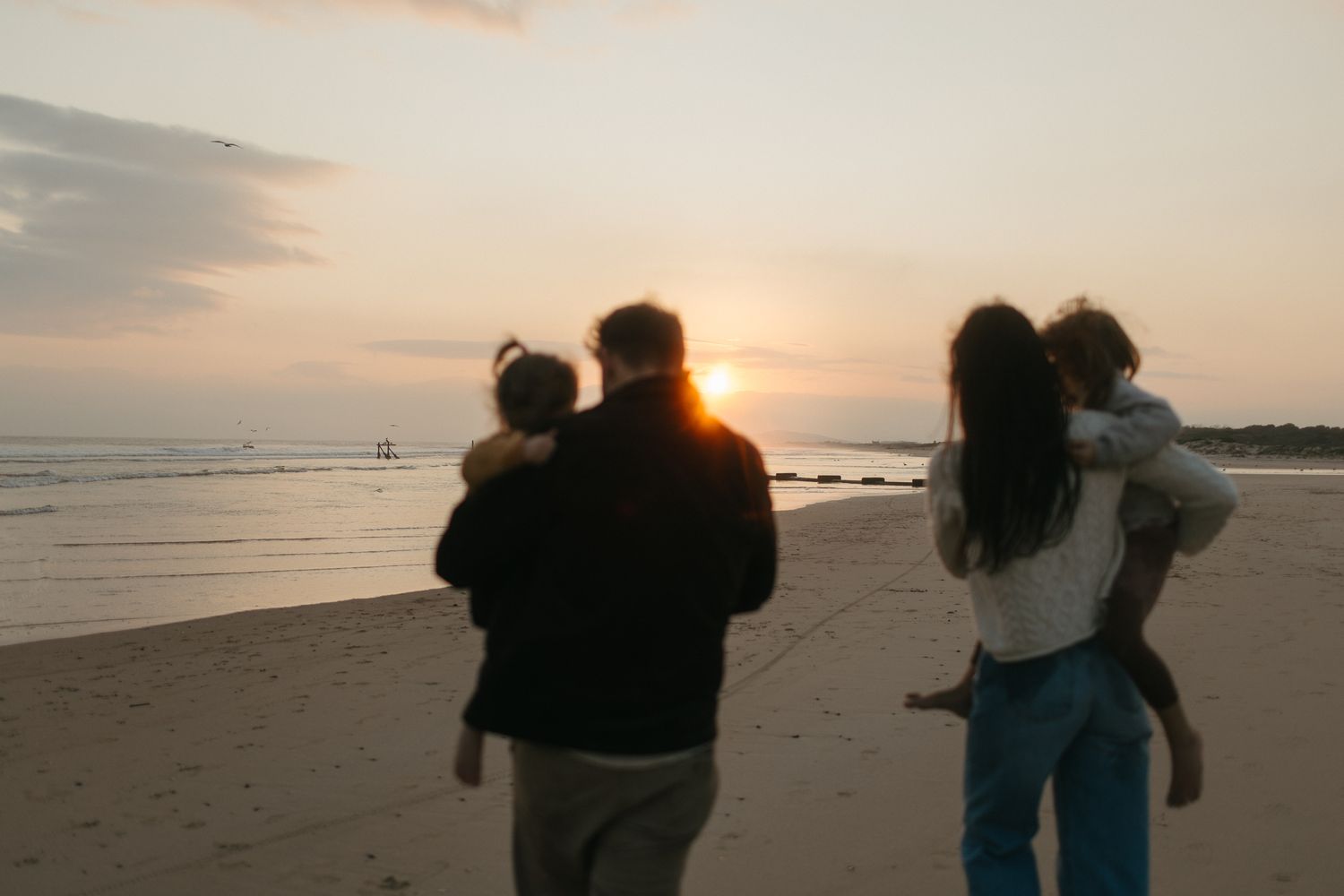 Family of four walking along the beach at sunset, parents carrying their two young children toward the glowing horizon.